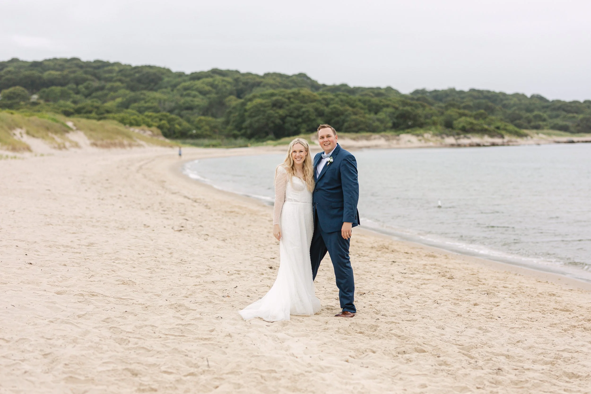 A bride and groom standing on a sandy beach with waves and green trees in the background. The bride is wearing a white wedding dress, and the groom is dressed in a blue suit with a bow tie. They are smiling and holding hands.