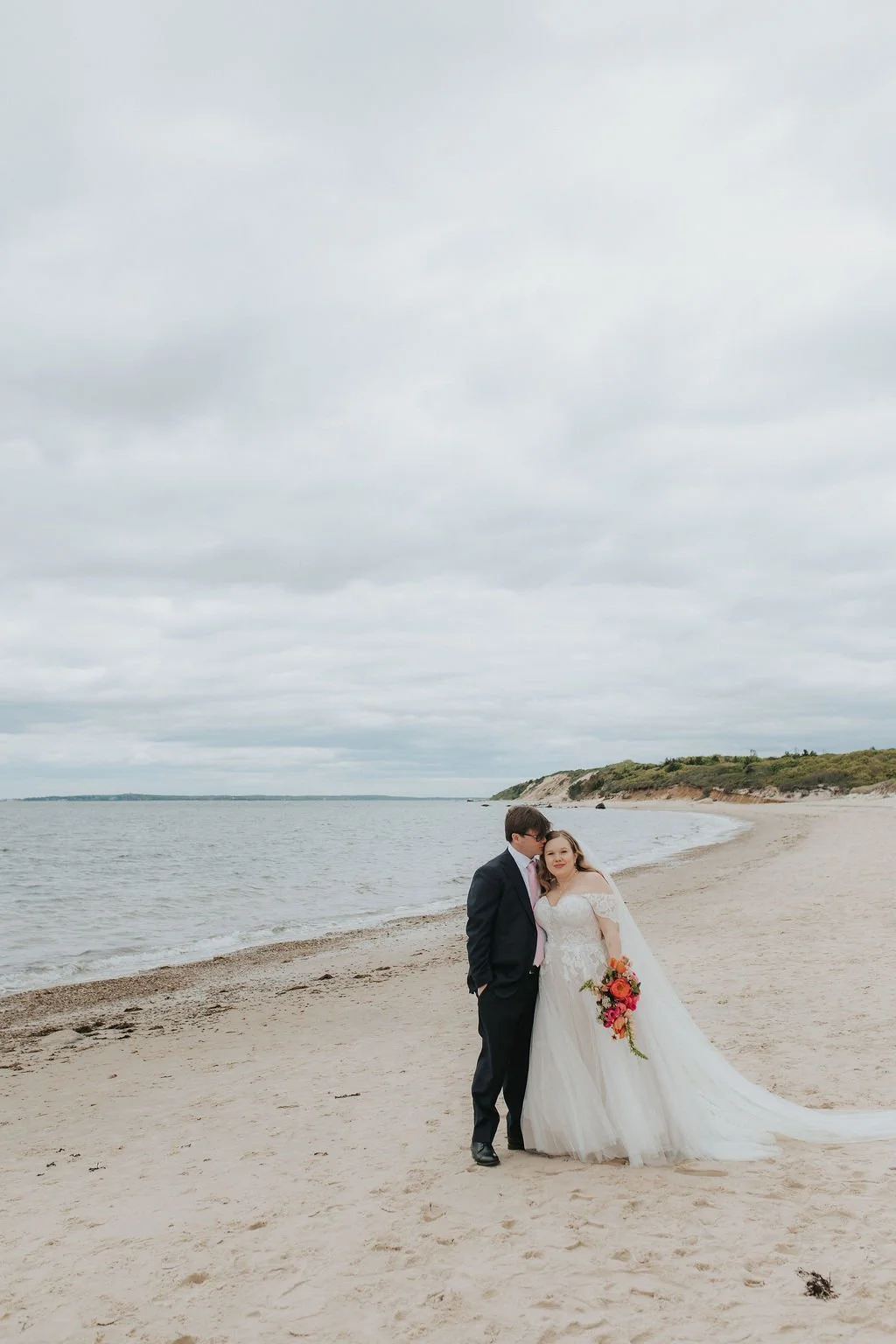 A bride and groom standing closely together on a beach at Lambert's Cove Inn on Martha's Vineyard, with the bride holding a bouquet of pink and orange flowers, under a cloudy sky.