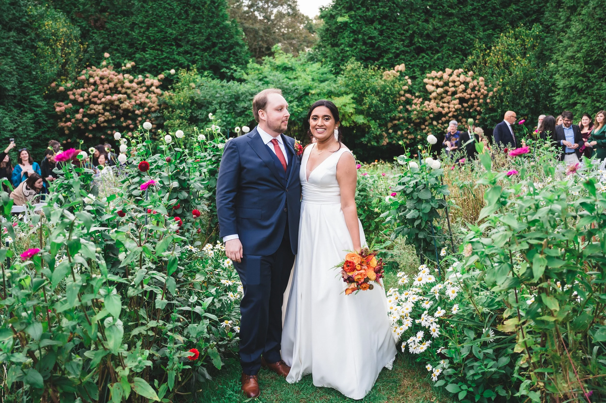 A bride and groom standing in a lush garden at Lambert's Cove Inn on Martha's Vineyard surrounded by flowers, with guests in the background at a wedding celebration.