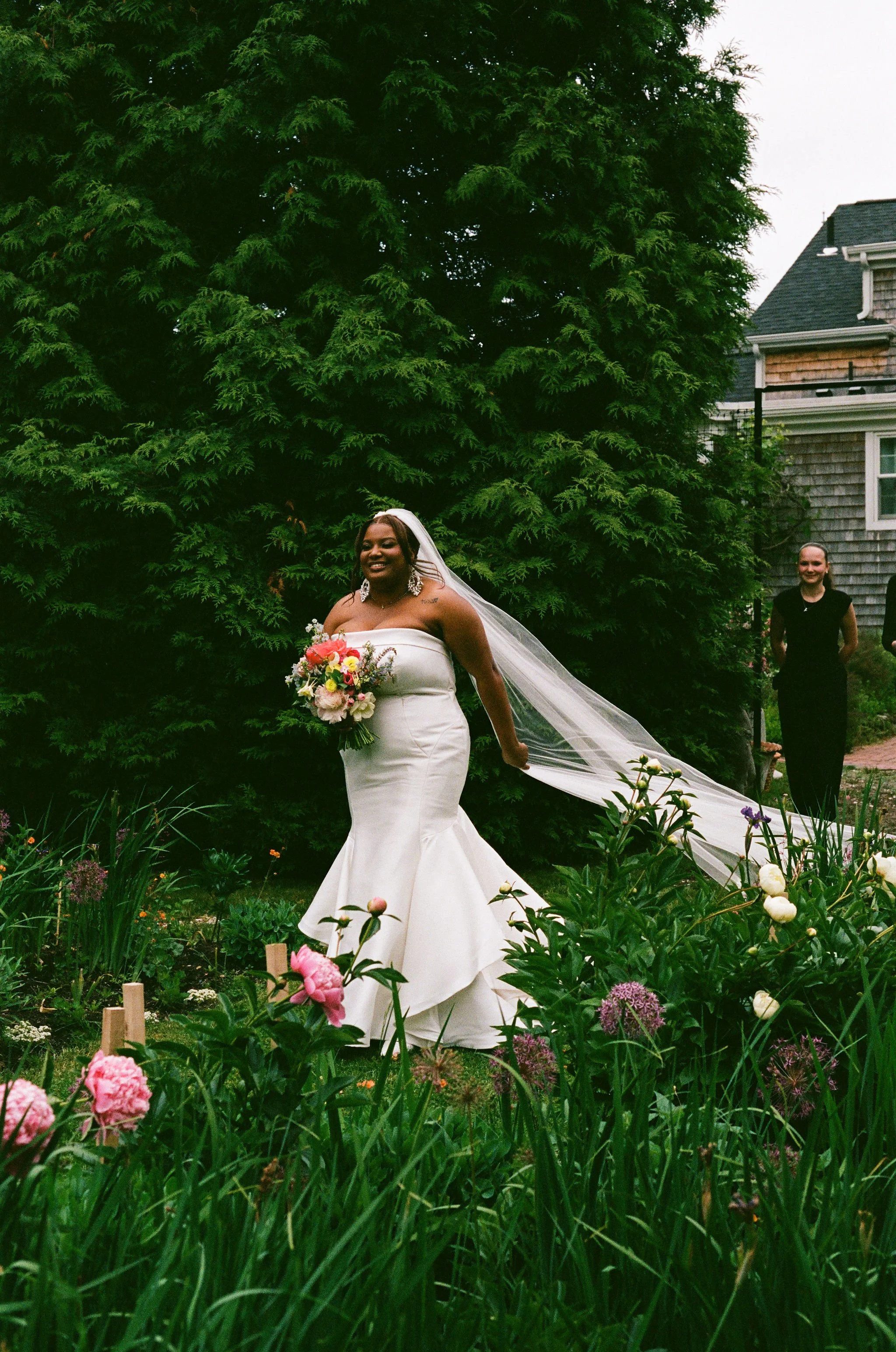A bride in a white dress holding a bouquet of flowers, standing in a garden with pink and white flowers in the foreground. There are people and a building with wooden shingles in the background at Lambert's Cove Inn on Martha's Vineyard.