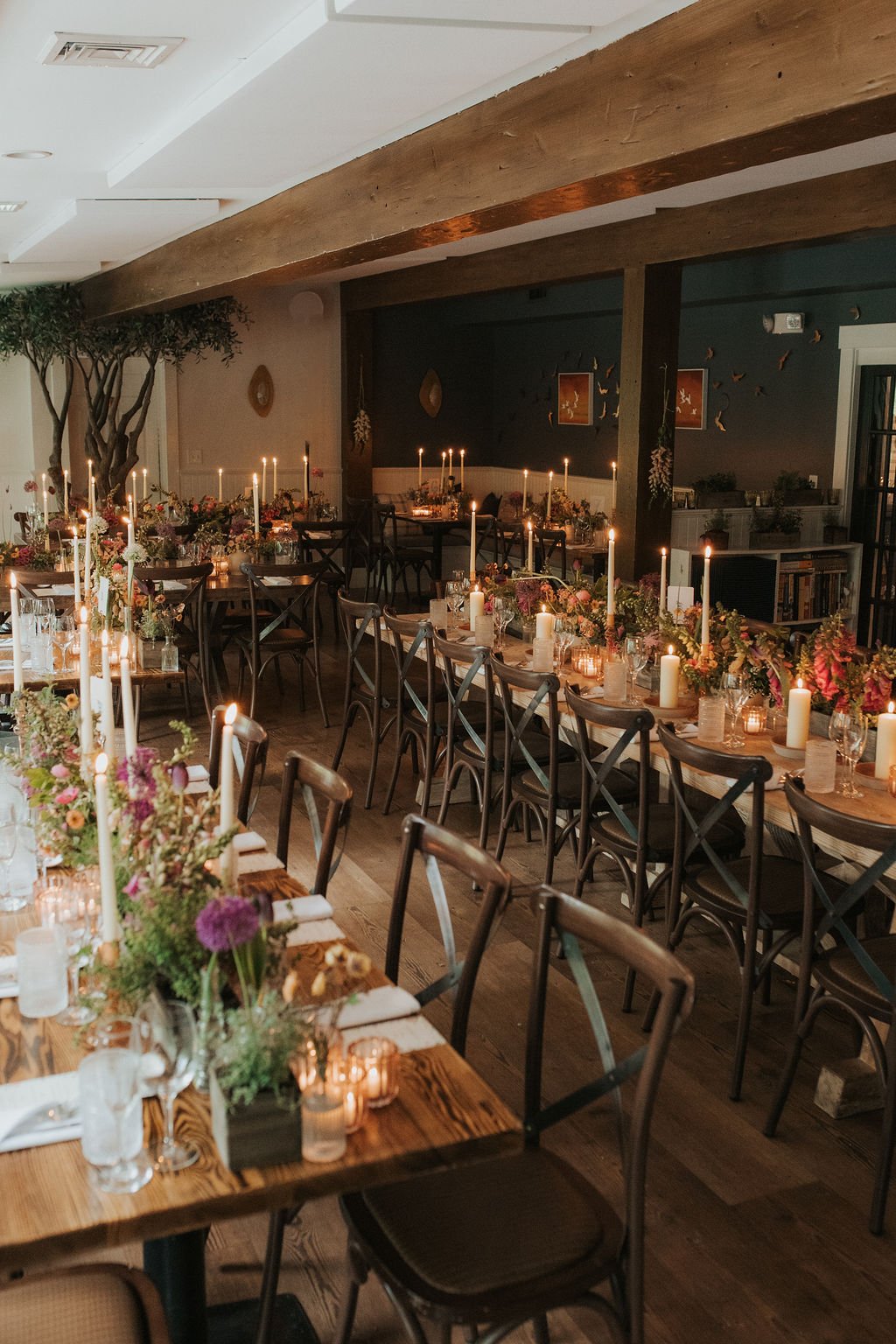 A cozy indoor dining area decorated for a celebration with long wooden tables, floral centerpieces, white candles, and soft candlelight lighting, alongside wooden chairs and minimal wall decor.