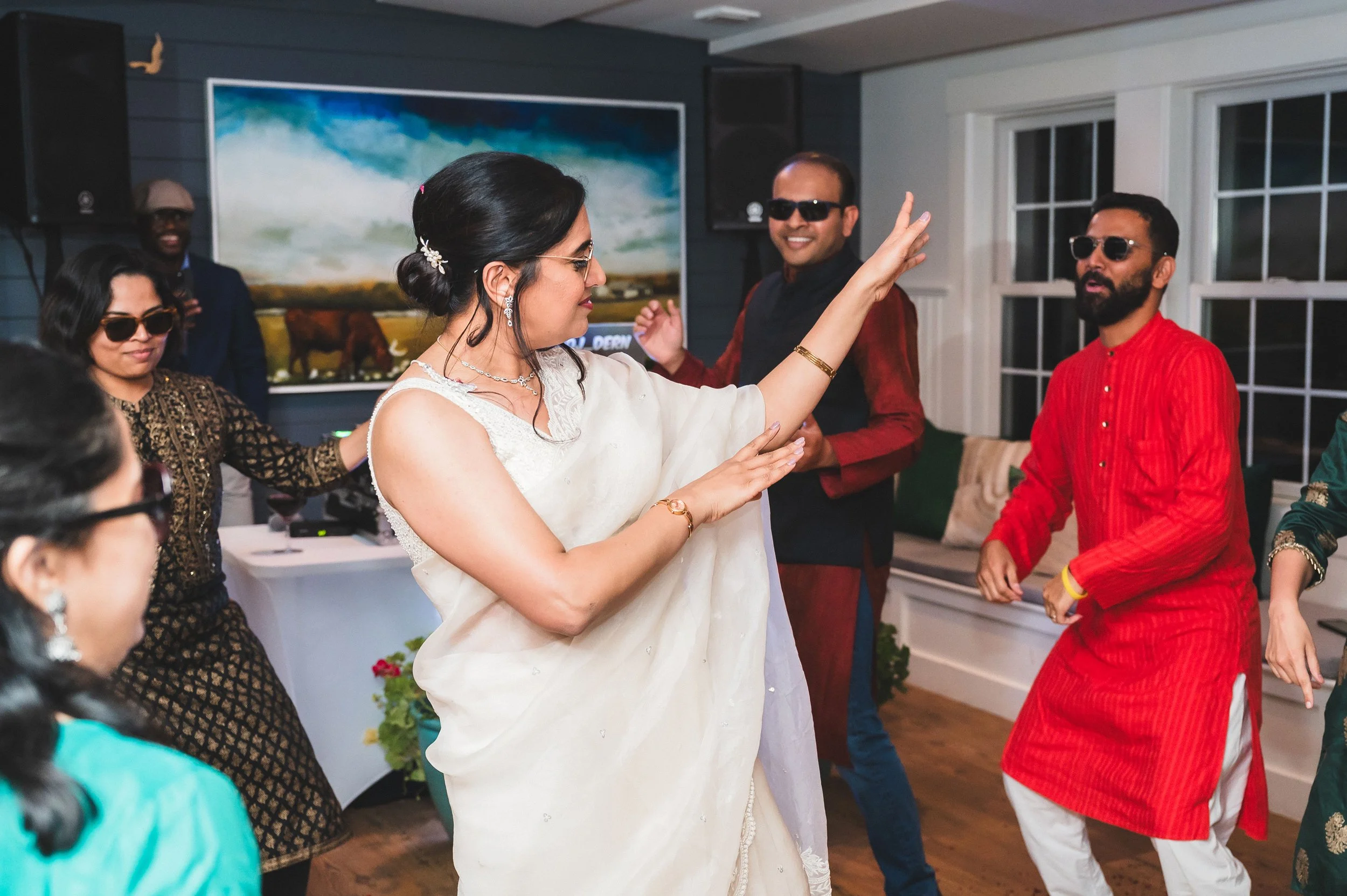 A woman in a white sari raising her hand as she dances at a gathering with several other people inside a house, some wearing sunglasses and colorful clothes, with a painting and large windows in the background at Lambert's Cove Inn.