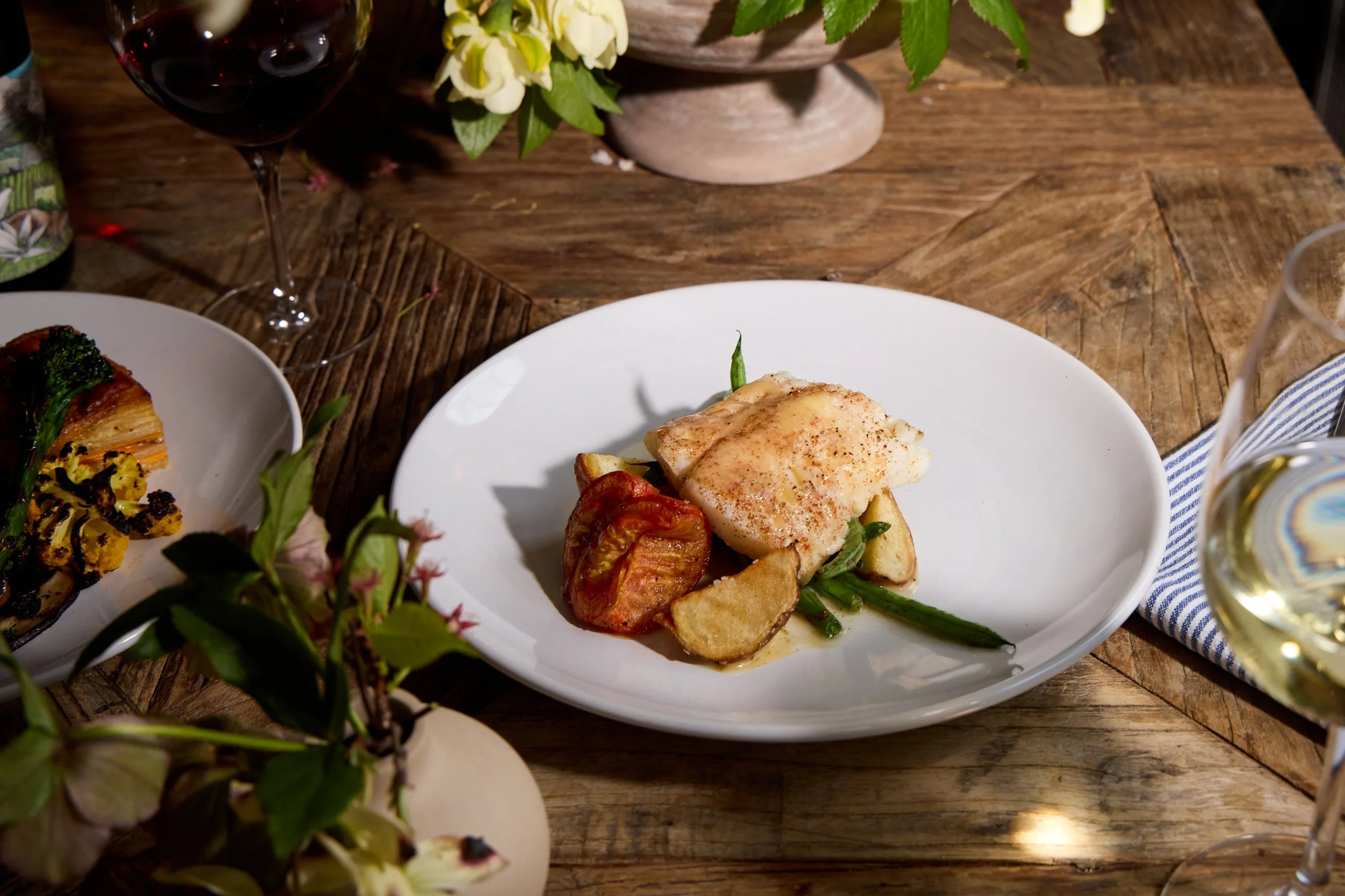 A white plate with a cooked fish fillet topped with spices, accompanied by roasted cherry tomatoes and green beans, set on a wooden table at Restaurant Foster at Lambert's Cove Inn on Martha's Vineyard.