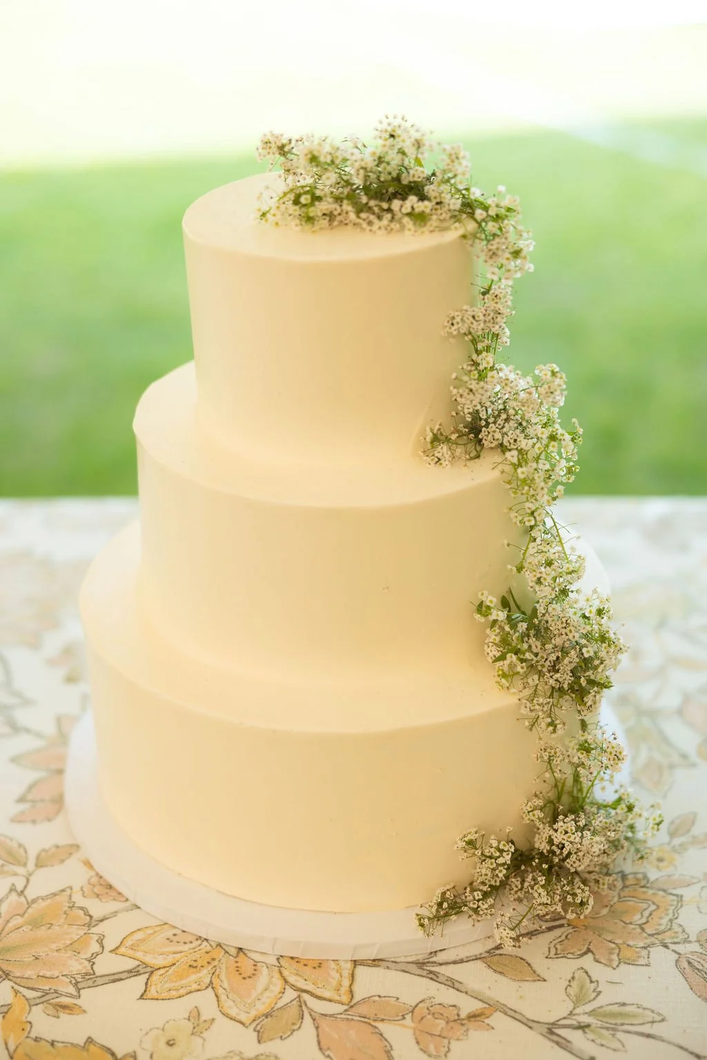 A three-tiered white wedding cake decorated with small white flowers cascading down one side, set on a floral tablecloth with a green outdoor background.
