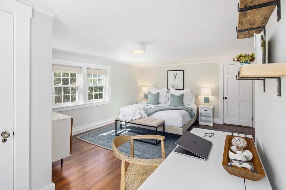 Bright bedroom with white walls and wooden floors, featuring a bed with white linens and gray pillows at Lambert's Cove Inn on Martha's Vineyard with two nightstands with lamps, a framed art piece of a deer above the bed, and a bench.