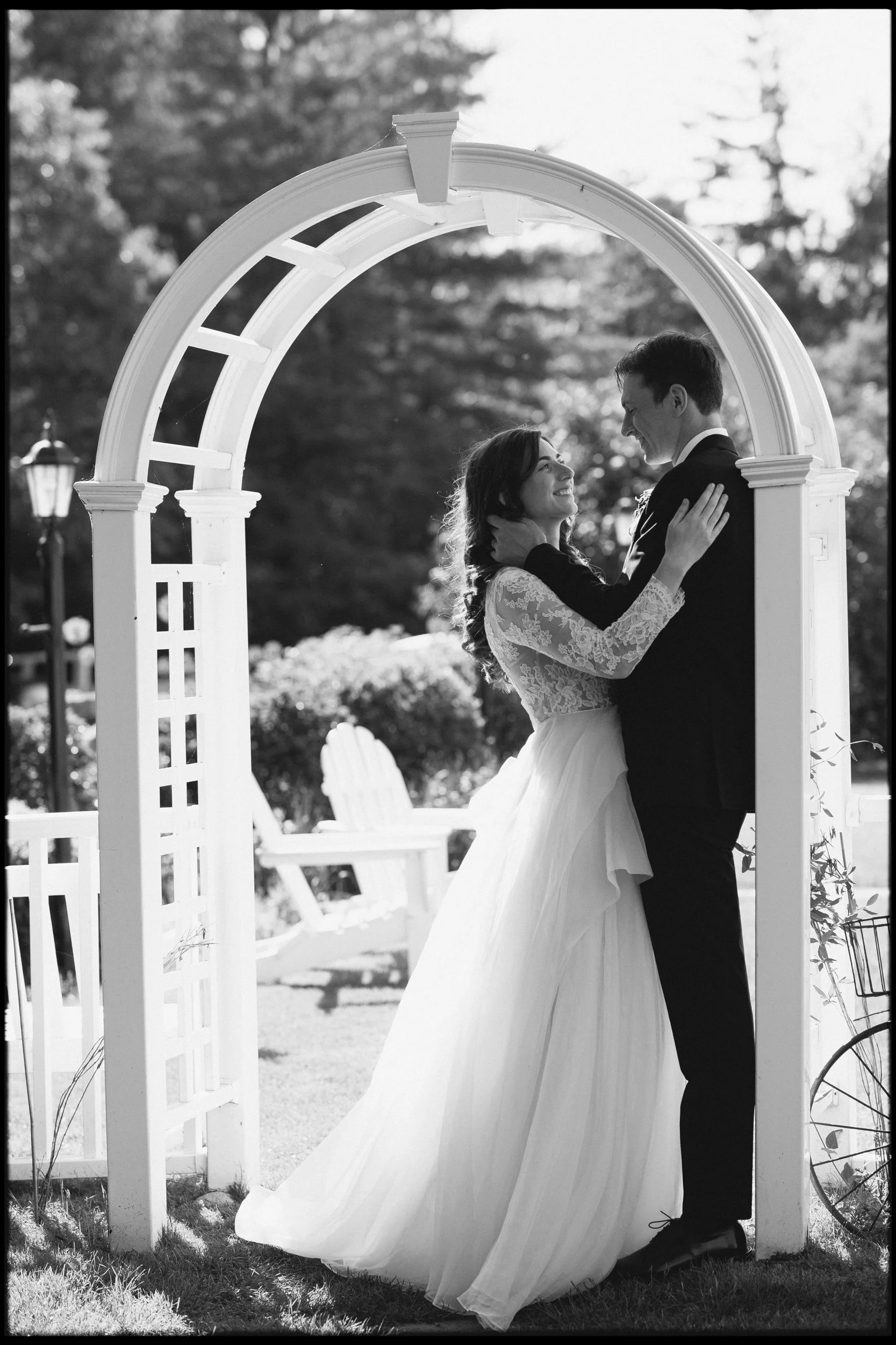 A black and white photo of a bride and groom smiling and holding each other under an archway, outdoors during a wedding at Lambert's Cove Inn on Martha's Vineyard.