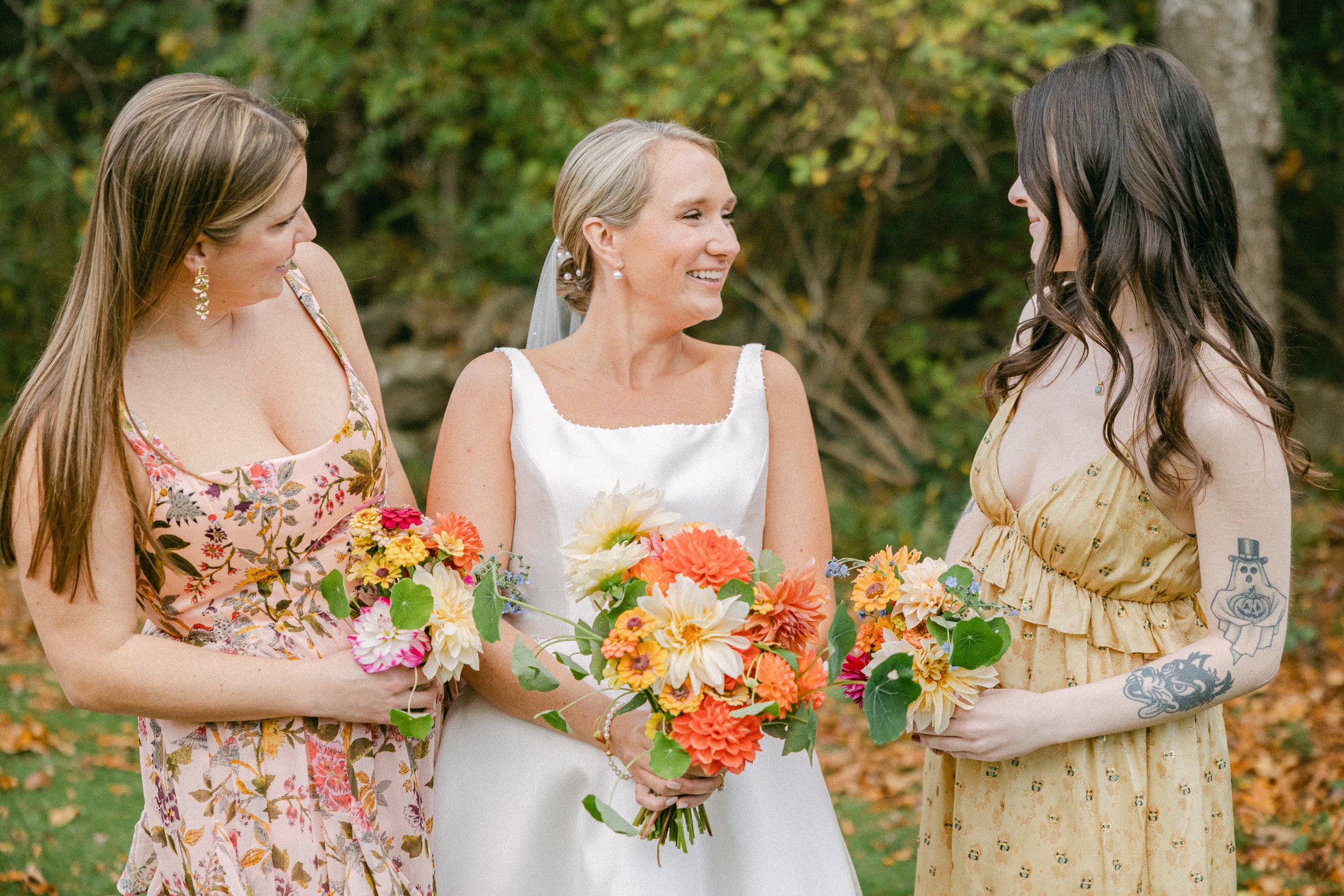 Three women standing together outdoors with trees and fallen leaves in the background, holding a bouquet of colorful flowers, dressed in summer attire, looking at each other and smiling during a wedding reception at Lambert's Cove Inn.