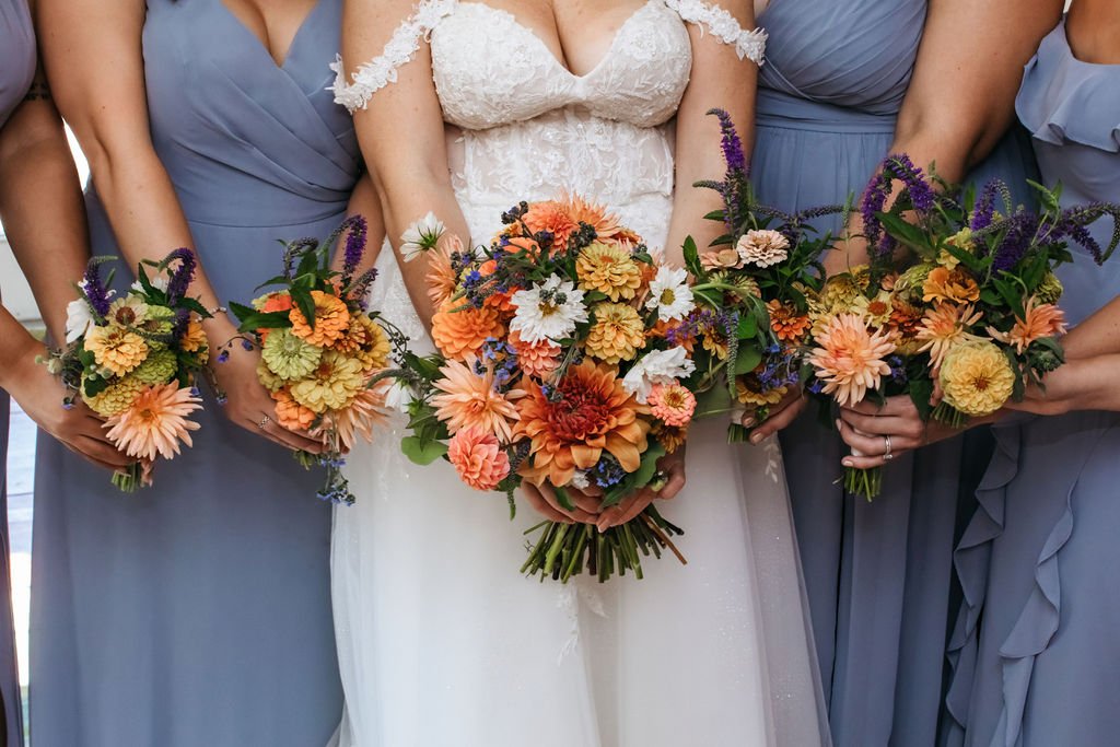 A bride in a white lace wedding dress holding a bouquet of orange, peach, white, and purple flowers, flanked by bridesmaids in blue dresses holding similar bouquets, getting ready for a wedding at Lambert's Cove Inn on Martha's Vineyard.