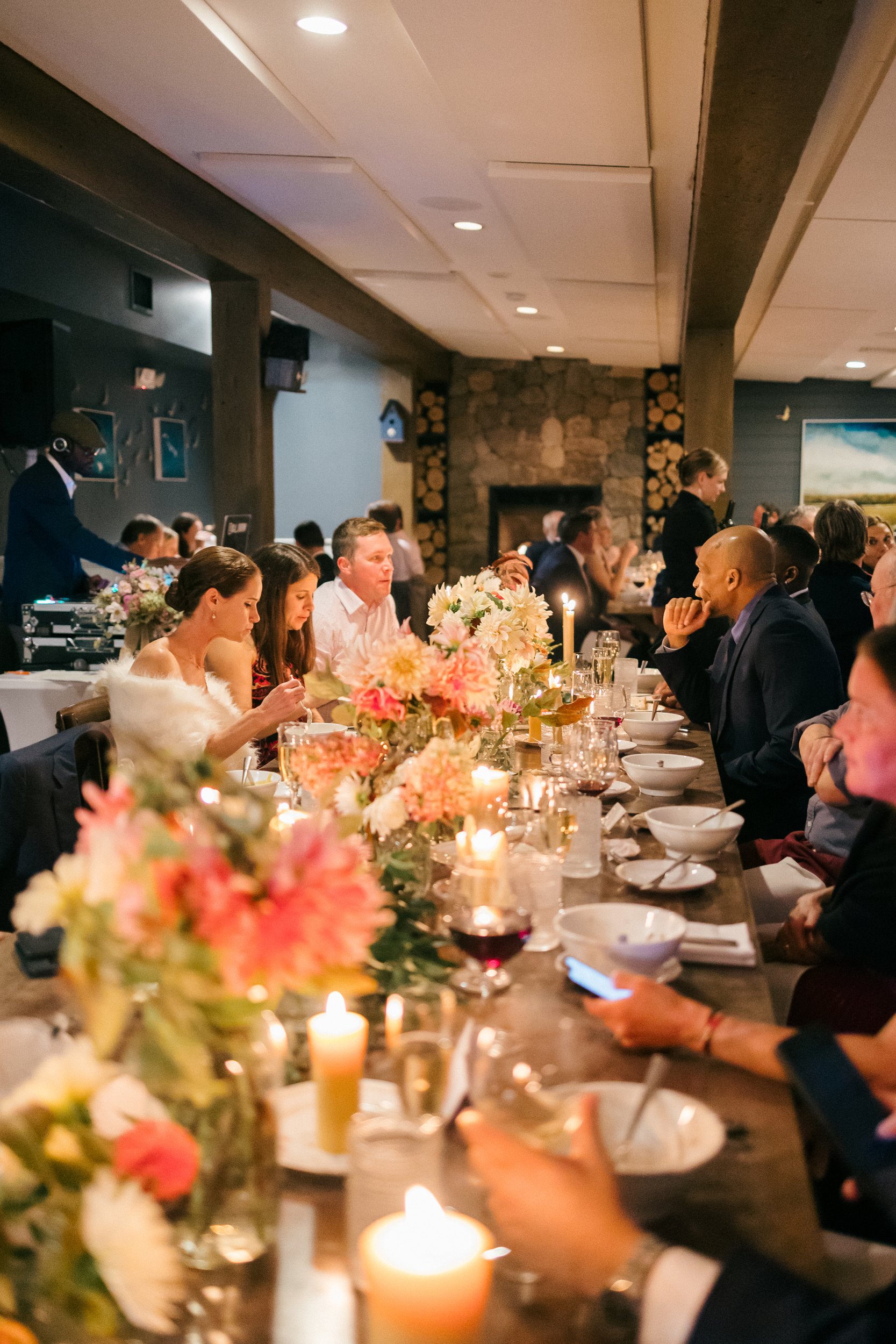 A group of people gathered around a long dinner table decorated with flowers and candles, in a warmly lit room with a stone fireplace in the background, at wedding reception at Lambert's Cove Inn on Martha's Vineyard.