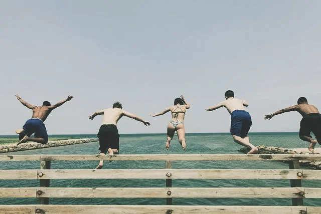 Five people jumping off a wooden pier into the ocean on a clear day.