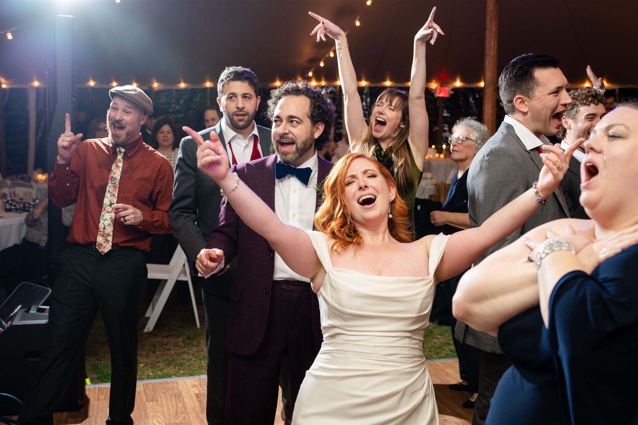 Group of people dancing and singing at a lively wedding reception at Lambert's Cove Inn on Martha's Vineyard with string lights, some with joyful expressions, others singing or dancing.