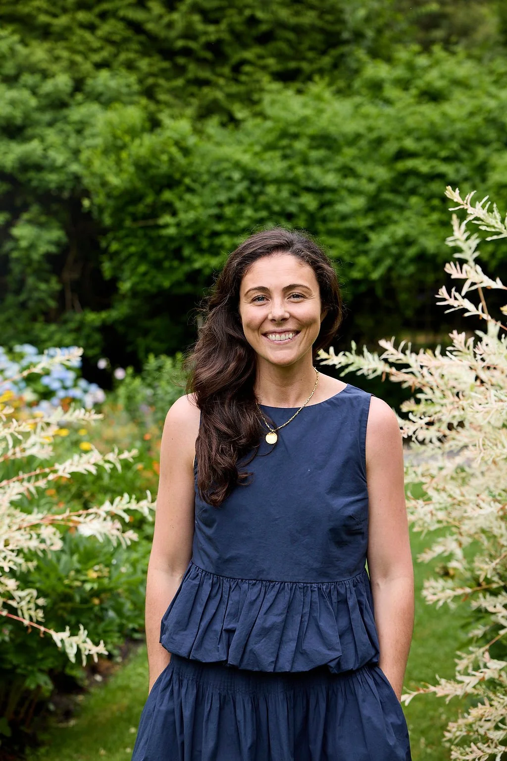 A woman, Olivia, Lambert's Cove Inn's Director of Rooms and Events, with long dark hair smiling, wearing a navy blue sleeveless dress and a gold necklace, standing outdoors in a lush garden with green foliage and flowering plants.