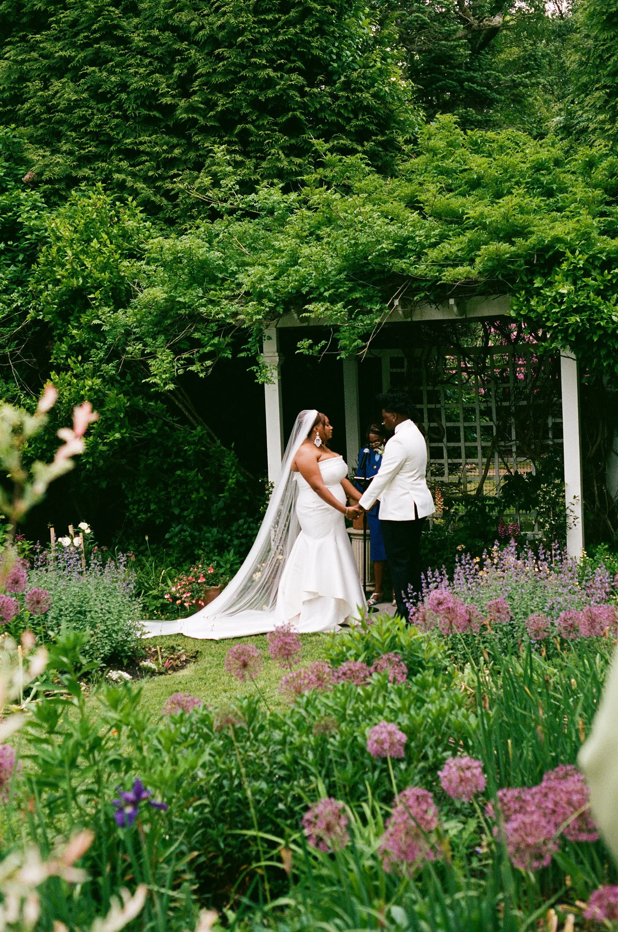 A bride and groom holding hands and exchanging vows outdoors under a canopy of green trees. The bride wears a white wedding dress and veil, and the groom a white suit. They are beside a white structure, surrounded by blooming purple and pink flowers.