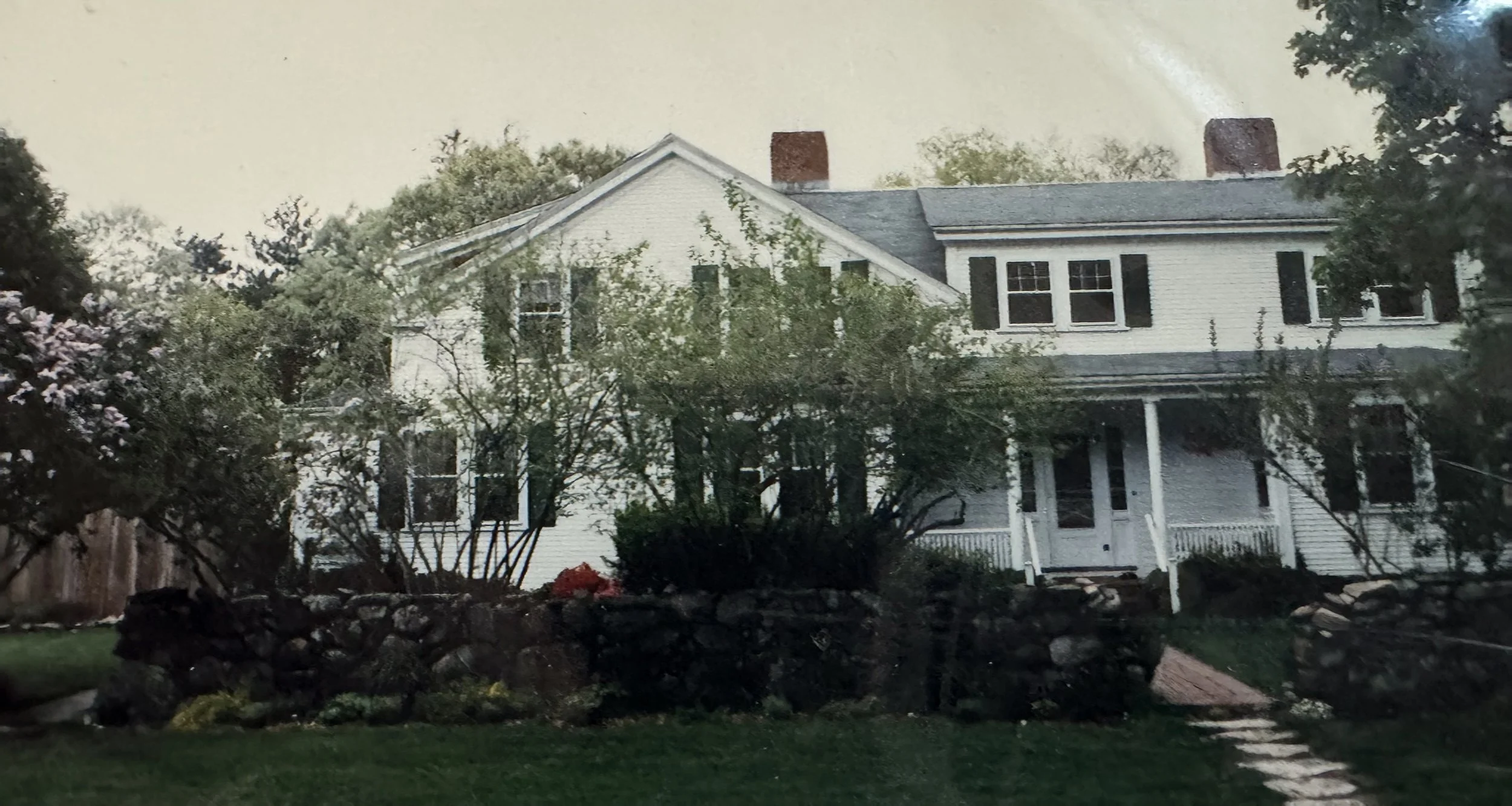 A white two-story house with black shutters, surrounded by trees and greenery, with a front porch and steps leading to the entrance.