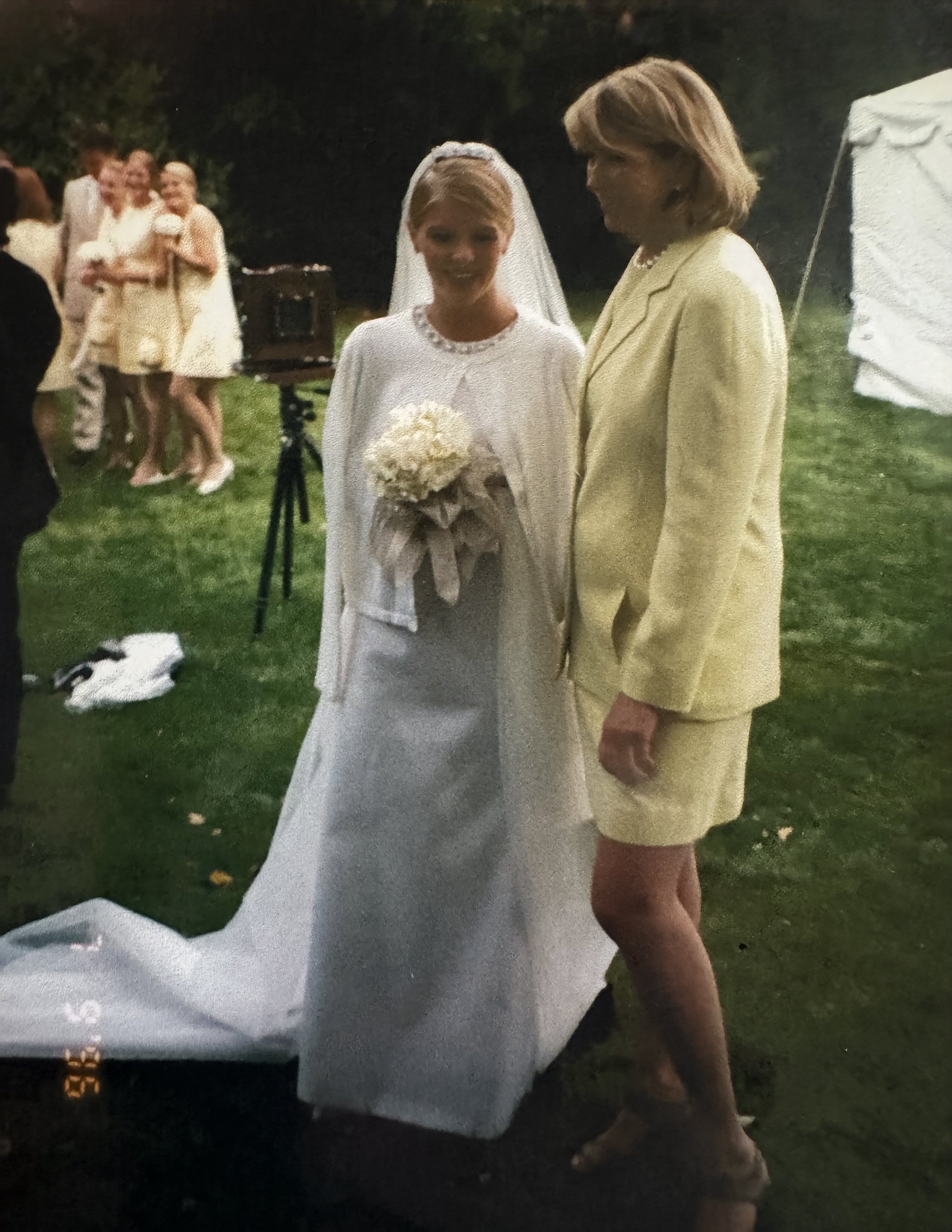 A bride in a white wedding dress and veil holding a bouquet, standing next to a woman in a yellow suit, at an outdoor wedding reception at night with guests in the background.
