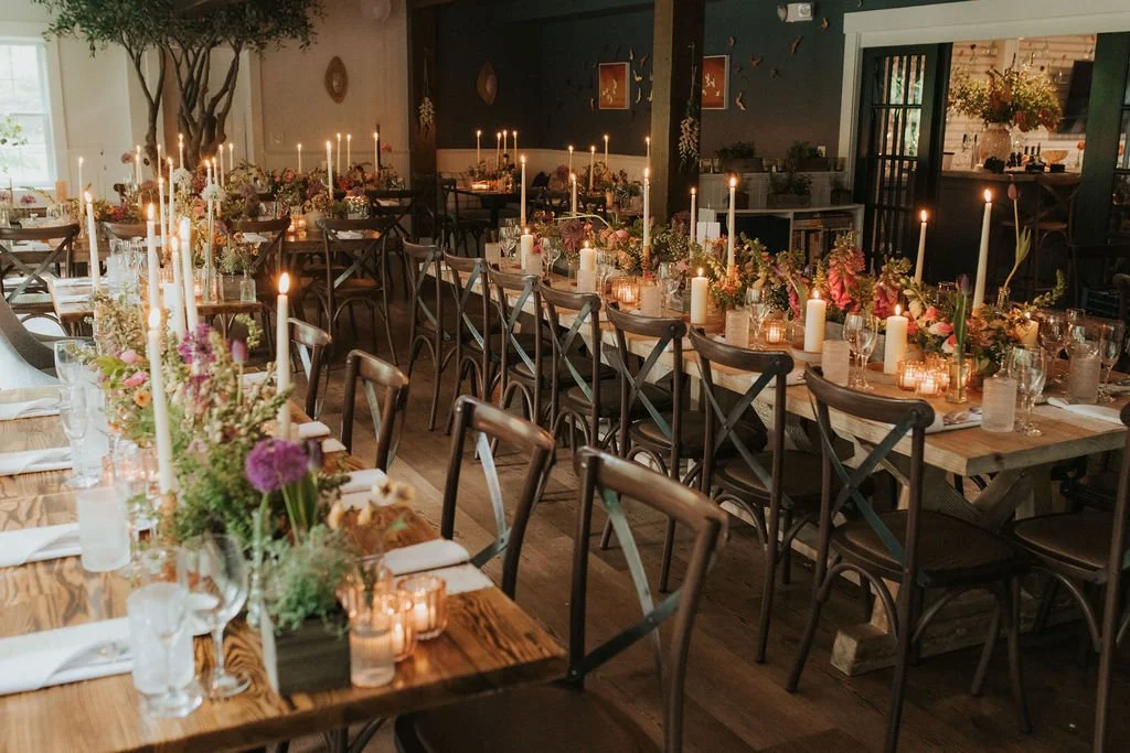 Long dining tables decorated with floral centerpieces and lit candles in a warmly lit event space.