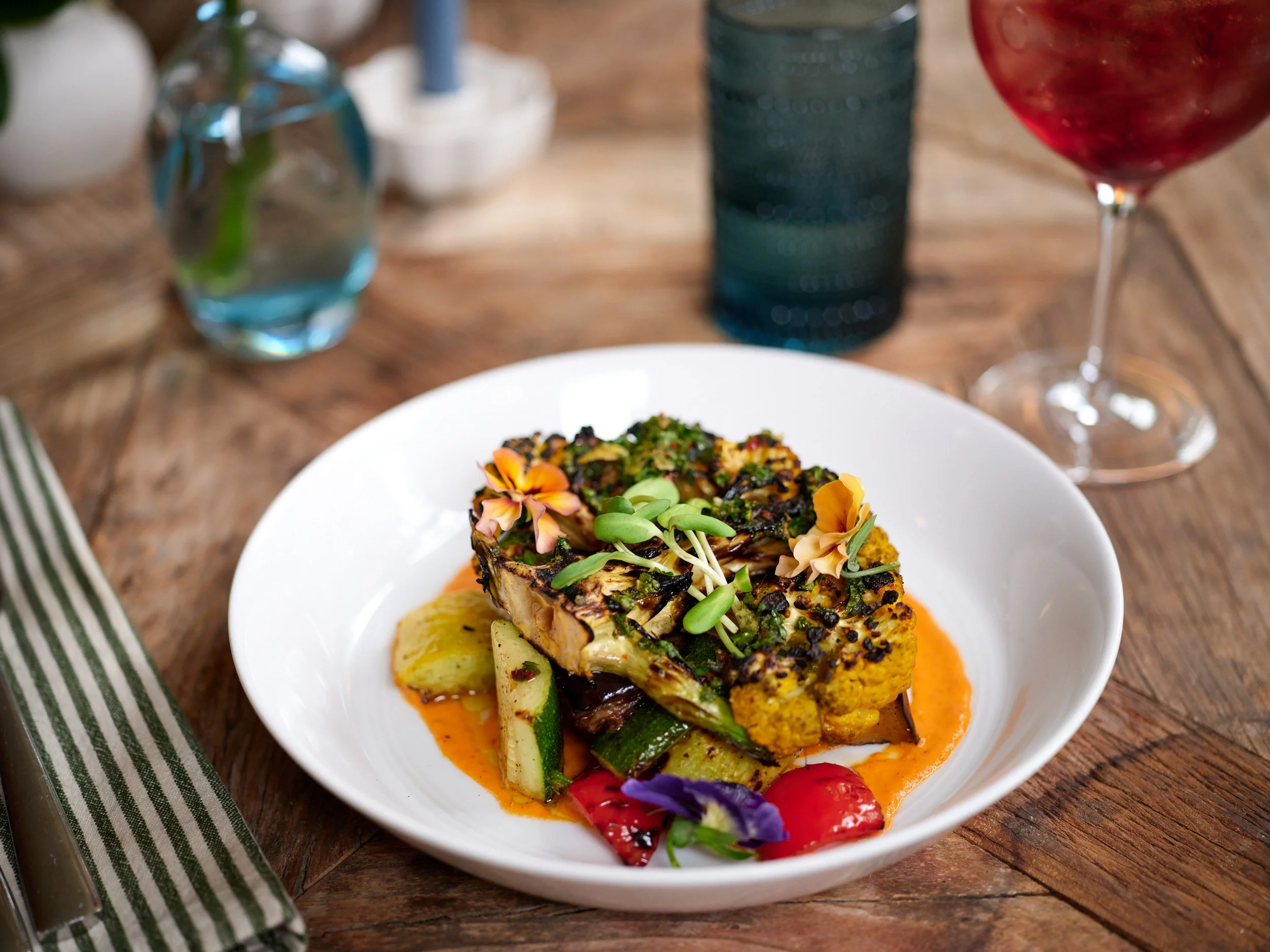 A plated dish featuring grilled fish topped with herbs and garnishes, served on a bed of roasted vegetables with colorful edible flowers, on a white plate at a rustic wooden table.