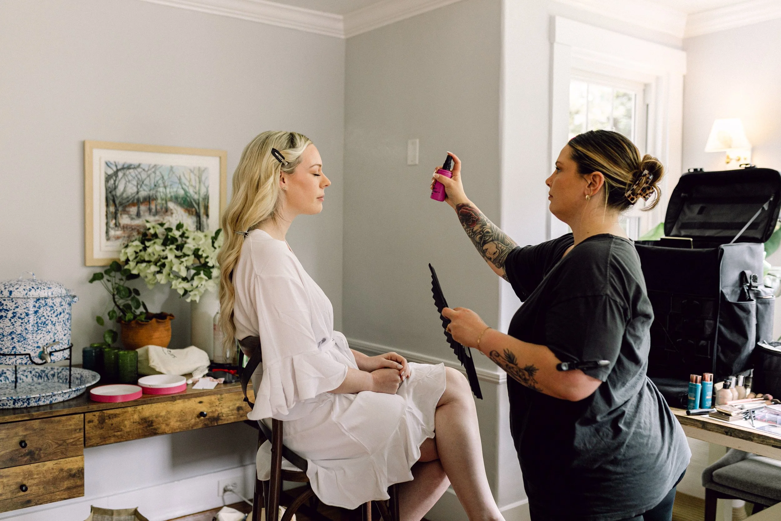 A woman with long blonde hair and a hairclip, sitting on a chair, looks straight ahead while another woman with tattoos and dark hair in a bun, wearing a black shirt, sprays makeup or hairspray on her face. The scene takes place in a well-lit room wi