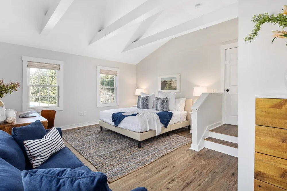 Bright bedroom with white walls, vaulted ceiling, two windows, a bed with white bedding and gray pillows, white nightstands with lamps, a blue sofa with striped pillows, a multicolored rug, and wooden flooring.