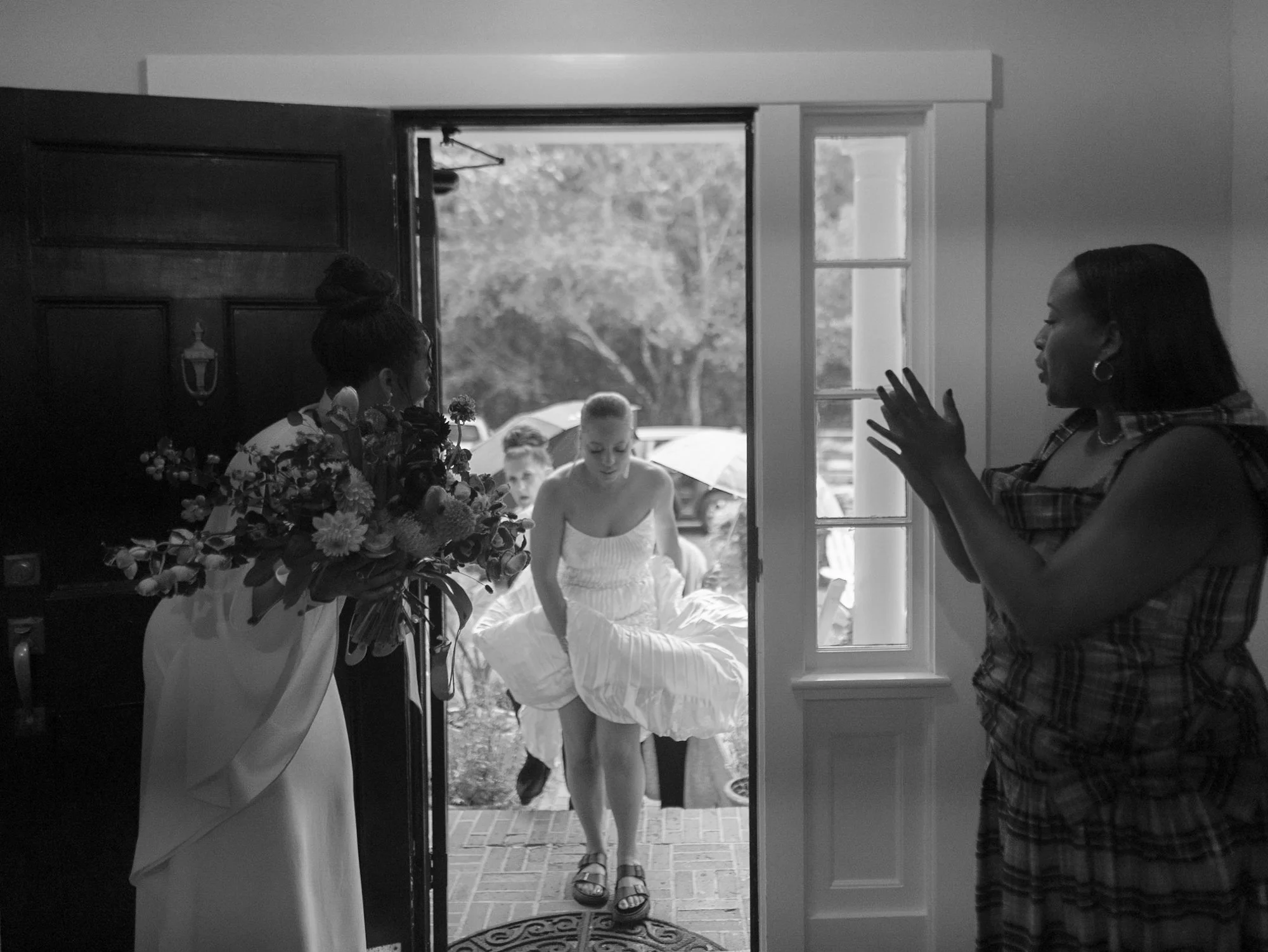 A girl in a white dress is stepping out of a house through a door, while an adult woman with shoulder-length hair and hoop earrings stands inside, facing her and gesturing with her hands. There are two children in the background outside, holding umbr