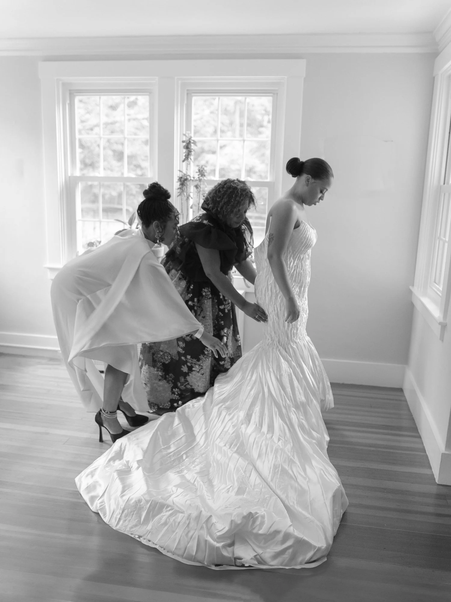 A bride getting ready with help from two women in a well-lit room with large windows at Lambert's Cove Inn on Martha's Vineyard.