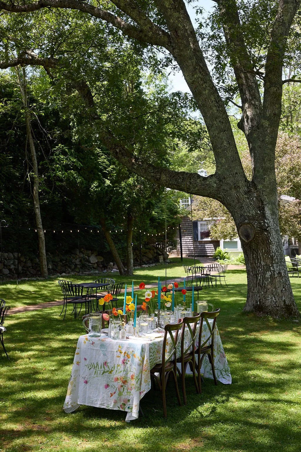An outdoor garden setting with a large tree, tables with floral tablecloths, colorful candles, and glasses, decorated for a celebration or event.