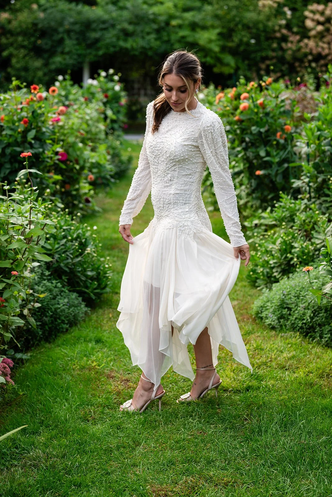 A woman in a white, long-sleeved, beaded dress standing on a grassy path in a garden with colorful blooming flowers and green trees.