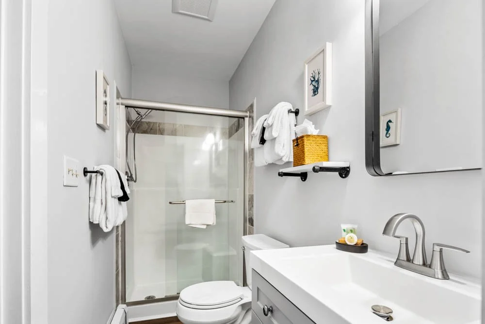 A small bathroom with a walk-in shower, white walls, a gray vanity with a white sink, a mirror, and framed artwork of sea creatures.
