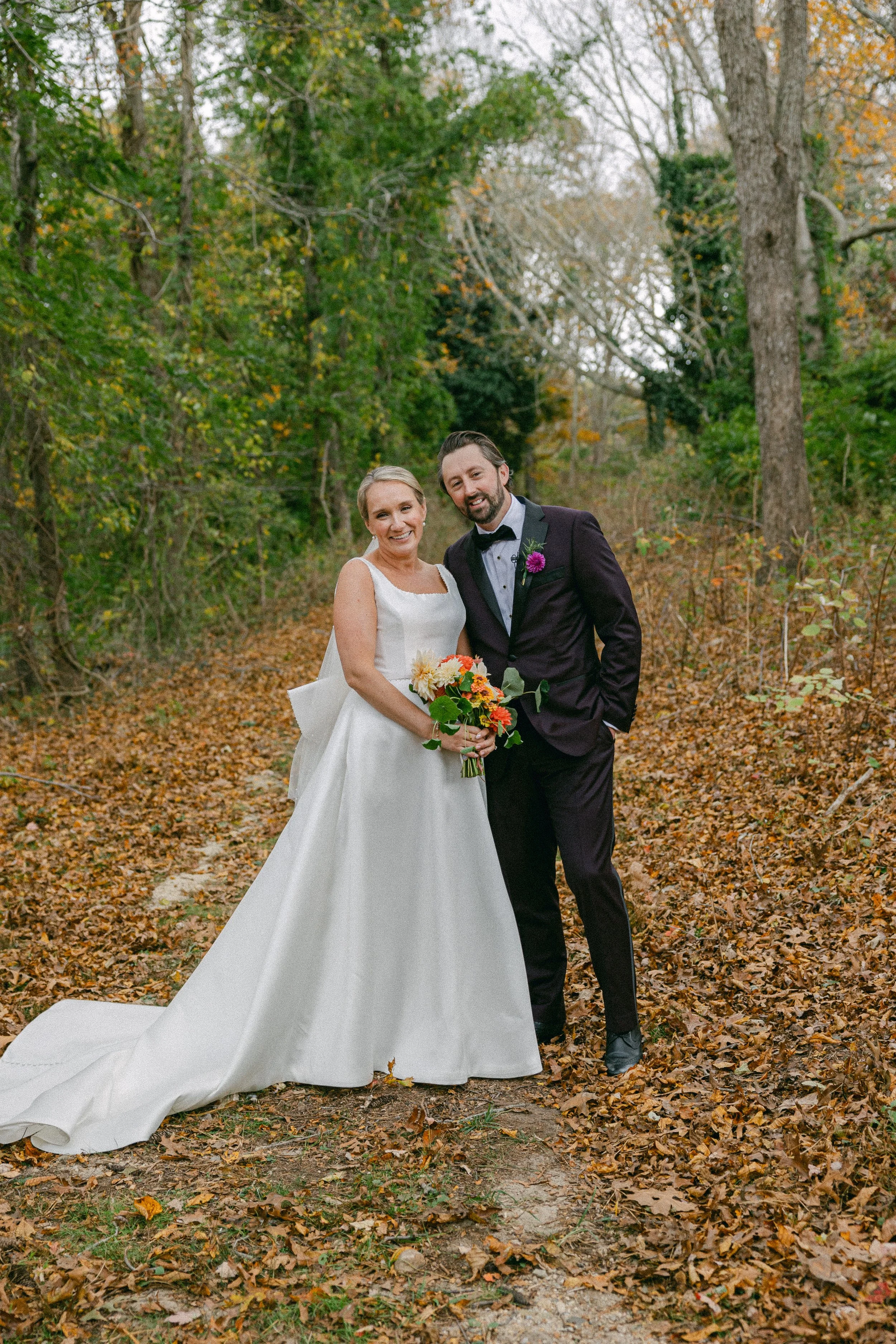 A bride and groom standing together in a wooded area with fall foliage, dressed in wedding attire, smiling and holding a bouquet of flowers during a wedding ceremony at Lambert's Cove Inn on Martha's Vineyard.