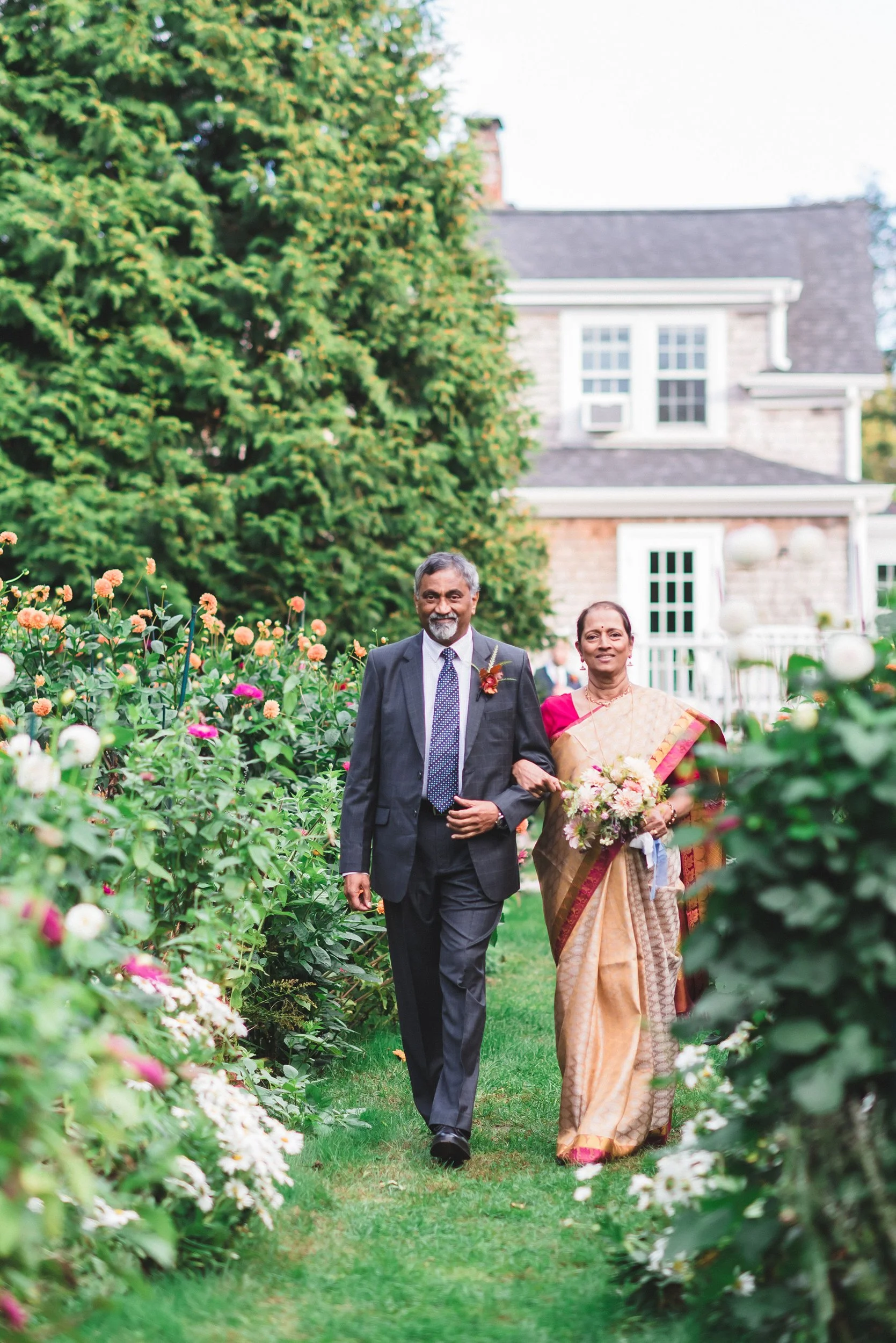 A couple walking arm in arm through a garden at Lambert's Cove Inn on Martha's Vineyard filled with blooming flowers, with a house and trees in the background.