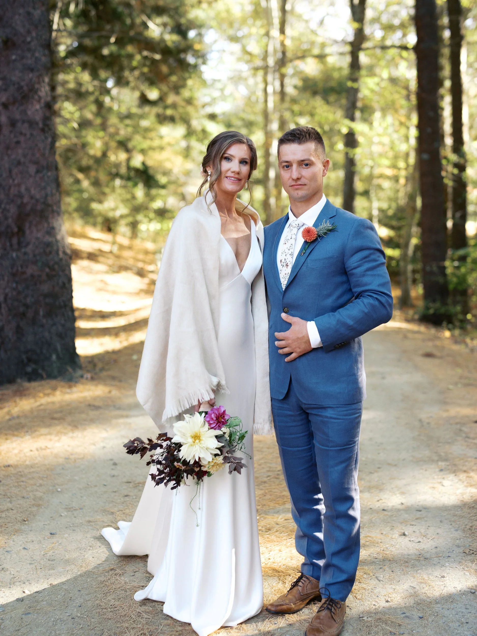 A bride in a white dress holding a bouquet of flowers. A groom in a blue suit with a boutonnière. They are in a forest setting during a wedding at Lambert's Cove Inn on Martha's Vineyard.
