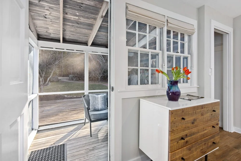 Interior view of a room with a wooden dresser topped with a purple vase holding orange tulips, next to a window and a sliding door leading outside to a deck with a chair, trees, and grass visible outside.