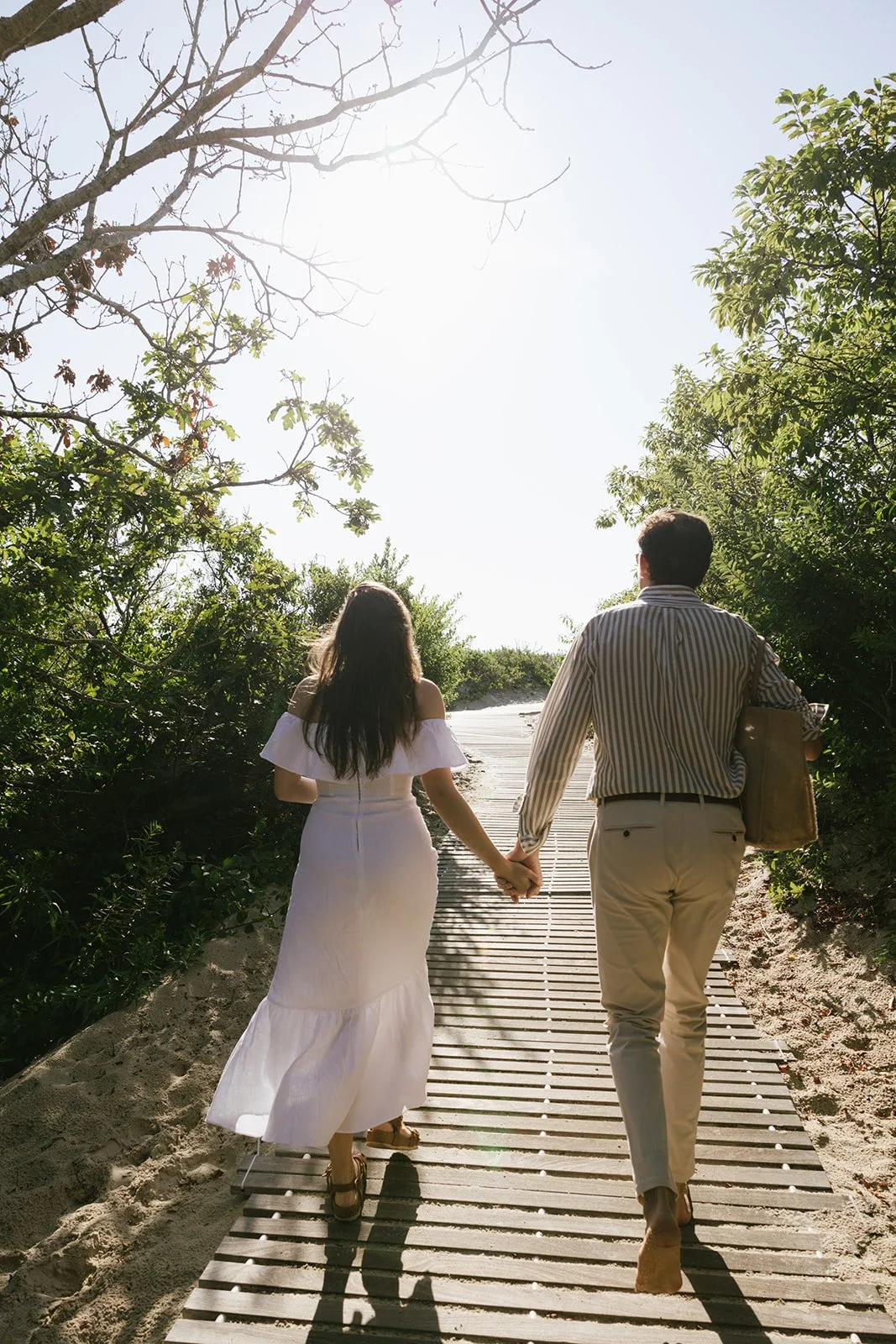 A man and woman holding hands walking on a wooden path through greenery, with sunlight overhead at Lambert's Cove Inn on Martha's Vineyard.