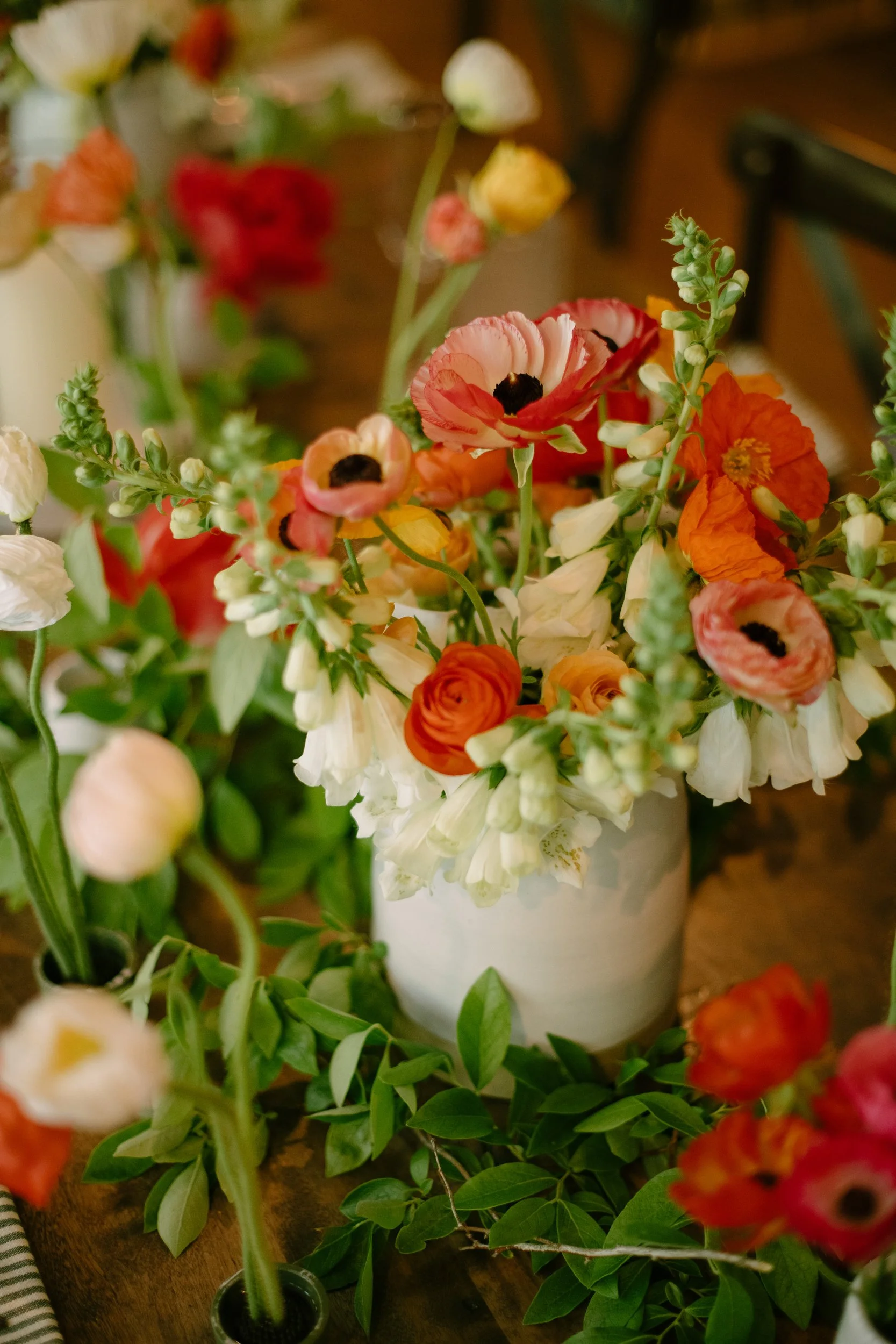 A variety of colorful fresh flowers in a white vase on a wooden table, with green leaves and stems surrounding them.