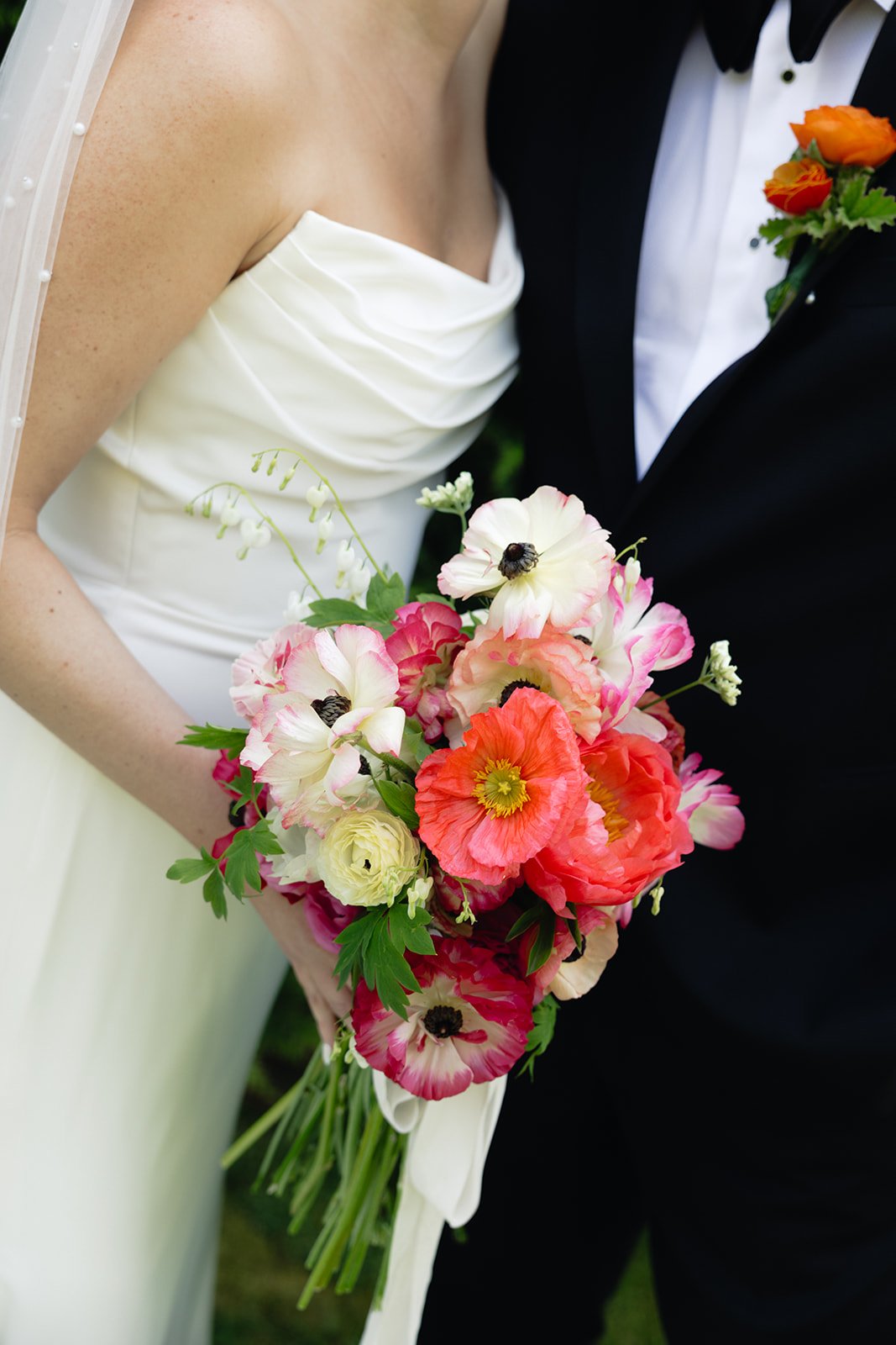 Close-up of a bride holding a bouquet with pink and white flowers, wearing a white wedding dress, and a groom in a black tuxedo with a floral boutonniere during a wedding at Lambert's Cove Inn on Martha's Vineyard.