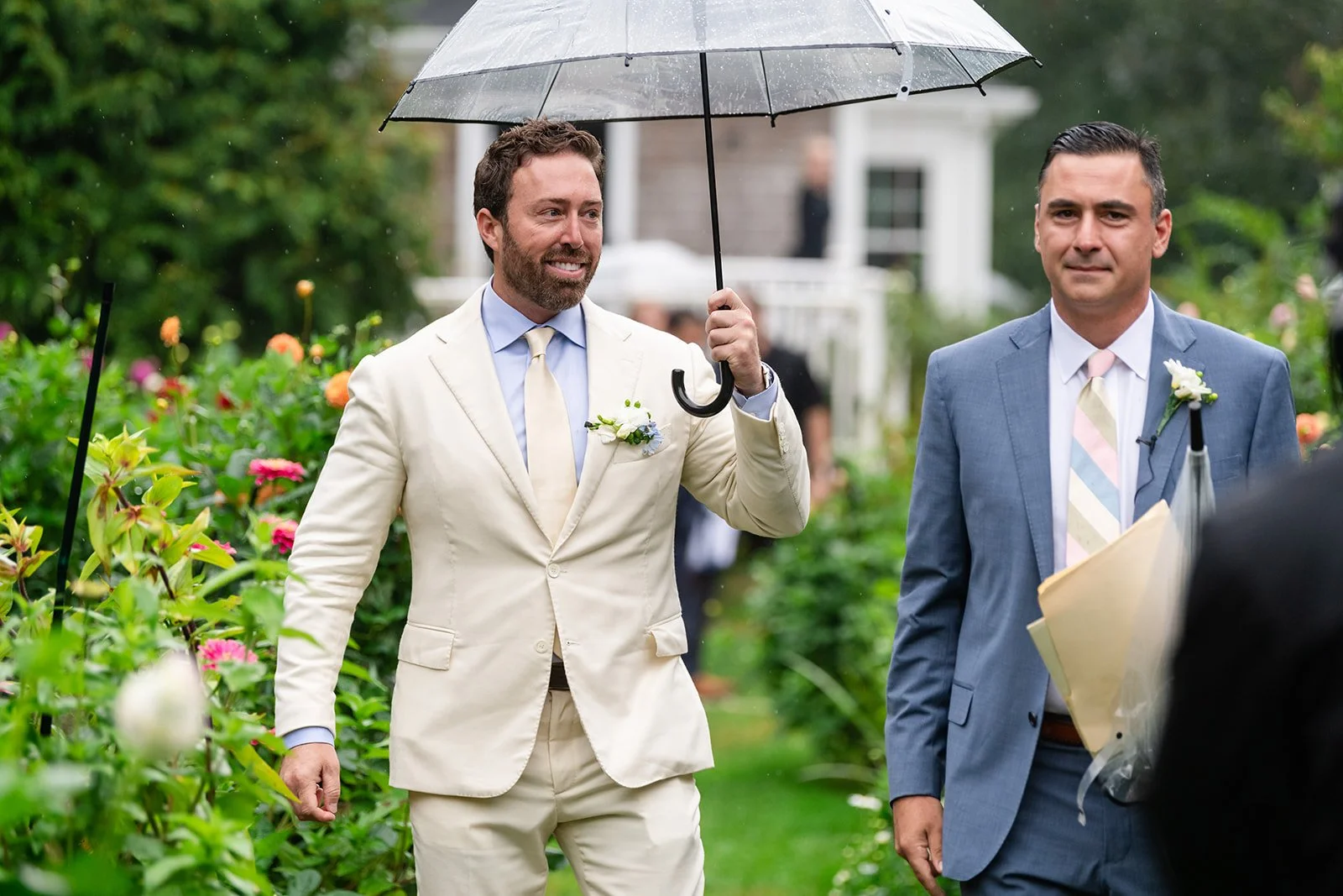 Two men dressed in suits are standing outdoors in a garden on a rainy day, holding umbrellas. One man, in a cream-colored suit, is smiling and holding a black umbrella. The other man, in a blue suit, looks serious. Both are wearing boutonnières and h