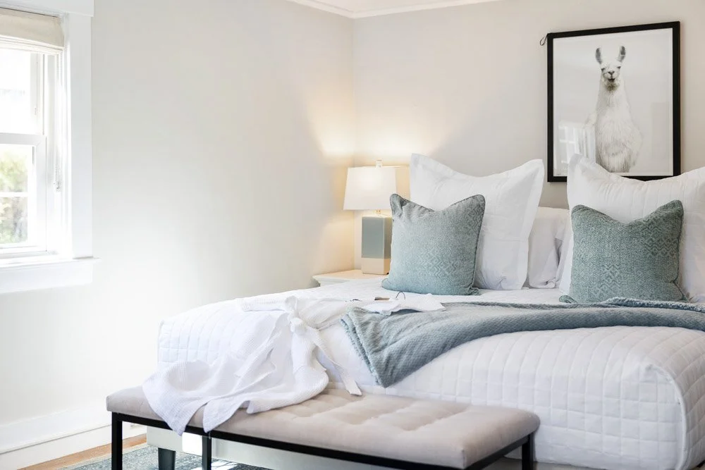 A neatly made bed with white and gray bedding, two gray pillows, a bedside lamp on a small white nightstand, a window with white blinds, and a framed picture of a llama on the wall.
