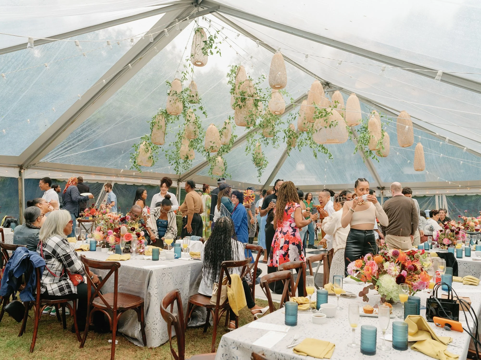 A lively gathering of diverse people at a wedding reception at Lambert's Cove Inn on Martha's Vineyard under a large tent, decorated with hanging woven lanterns and greenery, with tables set with colorful flowers, beige tablecloths, and tableware.
