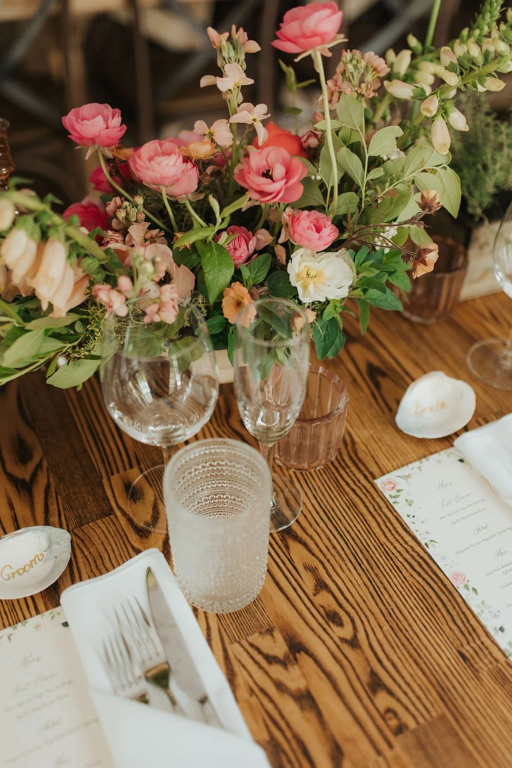 A floral centerpiece with pink, white, and peach flowers in a glass vase on a wooden table, set for a special occasion with glassware, utensils, a candle holder, and printed menus.