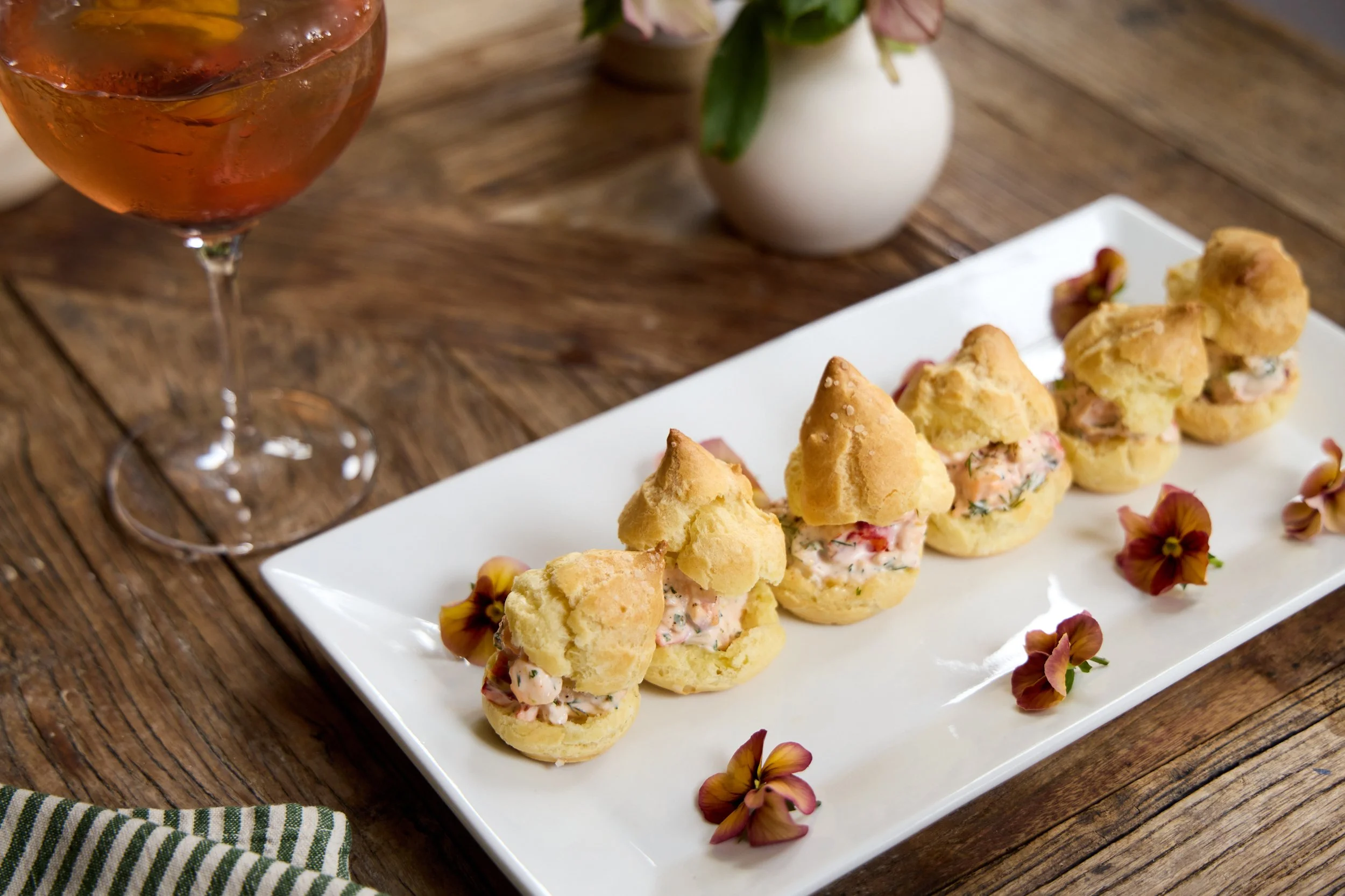 A white rectangular platter with puff pastry appetizers filled with a creamy seafood salad, garnished with edible flowers, on a wooden table next to a wine glass and a small white vase with pink flowers at Restaurant Foster on Martha's Vineyard.