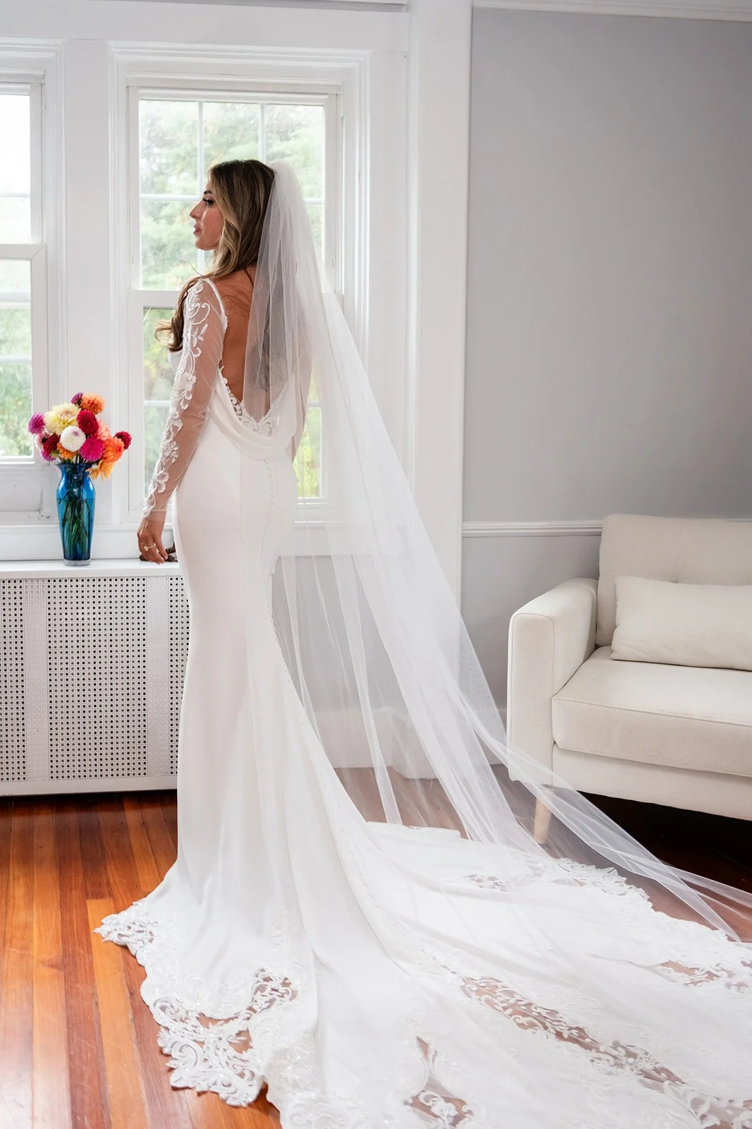 A bride in a white wedding dress with lace detailing, and a long veil, standing in a bright room near a window with a vase of colorful flowers on a radiator cover at Lambert's Cove Inn on Martha's Vineyard.