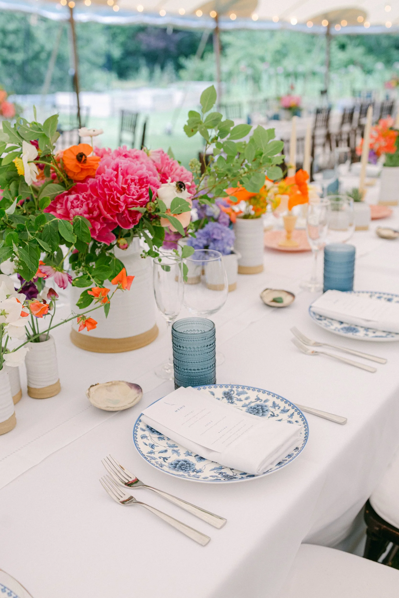 Elegant outdoor dining table decorated with colorful flowers in vases, set with blue and white floral plates, menus, forks, and glasses, under a tent with string lights during a wedding at Lambert's Cove Inn on Martha's Vineyard.