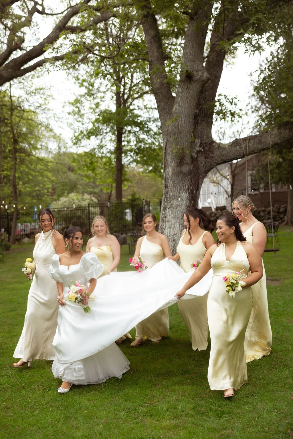 A bride and six bridesmaids in cream-colored dresses are outdoors on a grassy area, holding up the train of the bride's white wedding gown. They are smiling and holding small bouquets of flowers, with a large tree visible at Lambert's Cove Inn.