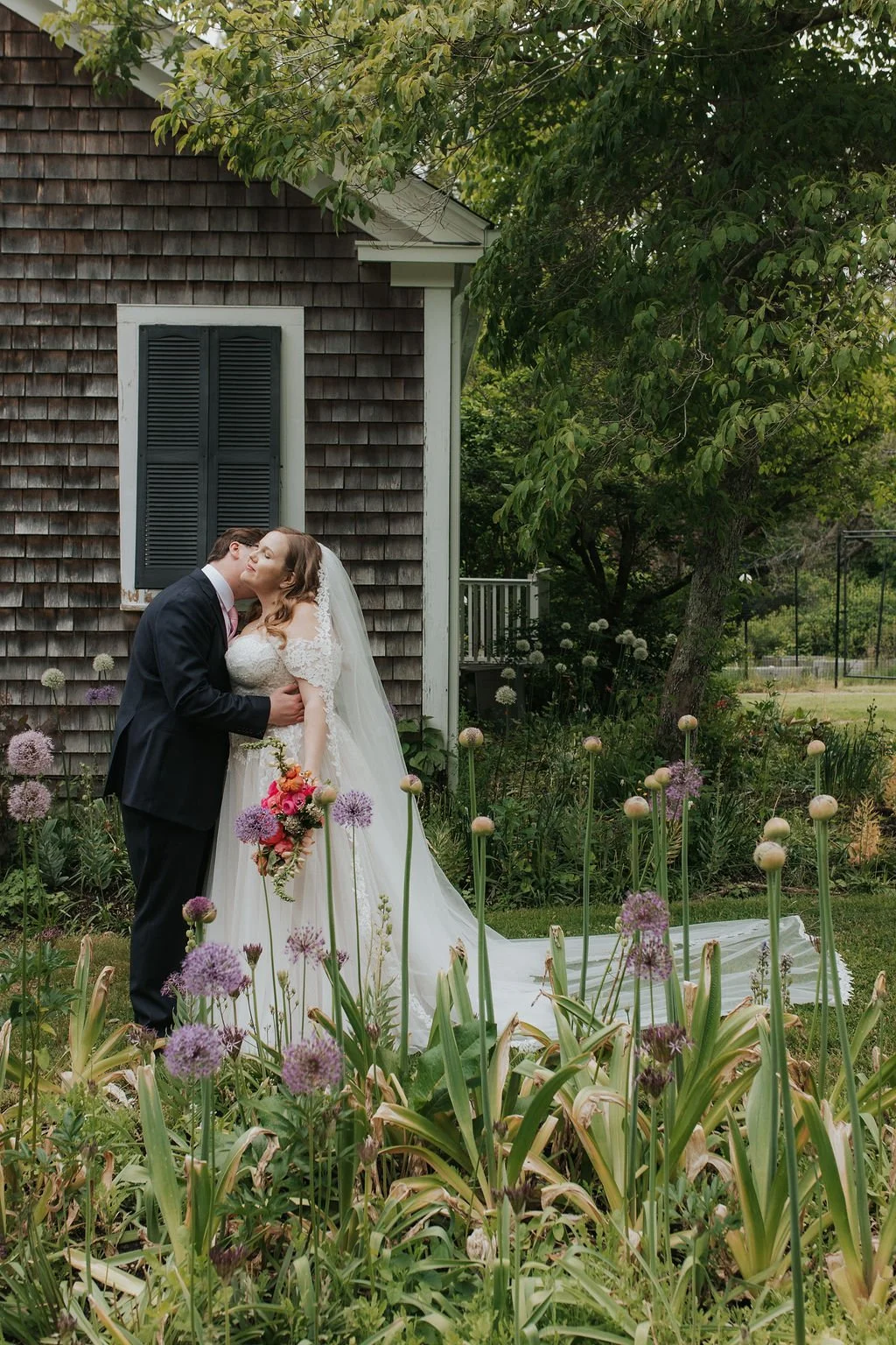 A bride and groom share a kiss in a garden at Lambert's Cove Inn on Martha's Vineyard, with purple flowers, next to a weathered wooden house with closed dark shutters.