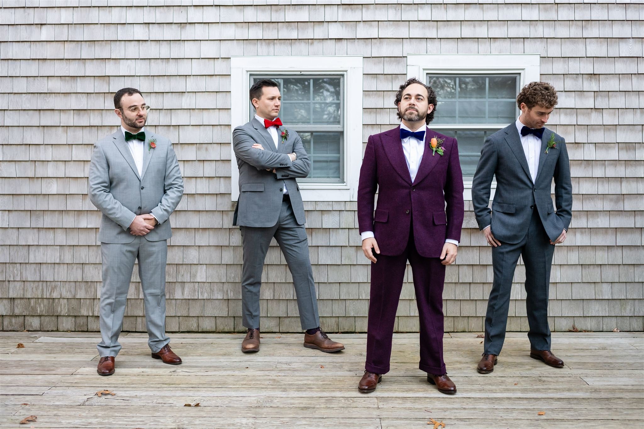 Four men dressed in suits with bow ties, standing outdoors in front of a house with wooden siding. The man in the center wears a purple suit and is looking upward, while the others wear gray and are looking in different directions.