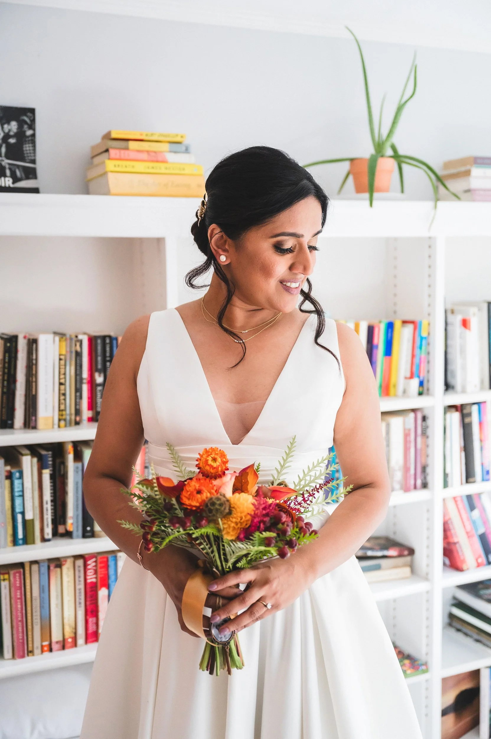 A woman in a white dress holding a colorful bouquet of flowers, standing in front of a bookshelf with books and a potted plant at Lambert's Cove Inn on Martha's Vineyard.