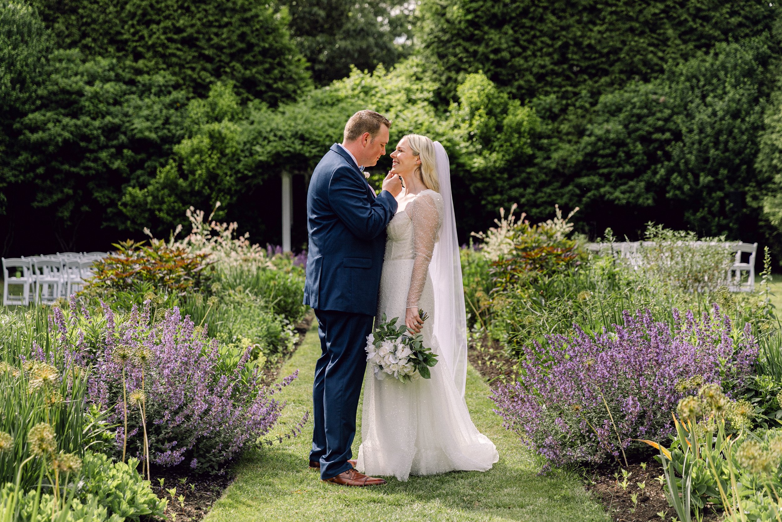 A bride and groom standing close together in a garden with purple flowers, gazing at each other during their wedding at Lambert's Cove Inn on Martha's Vineyard.