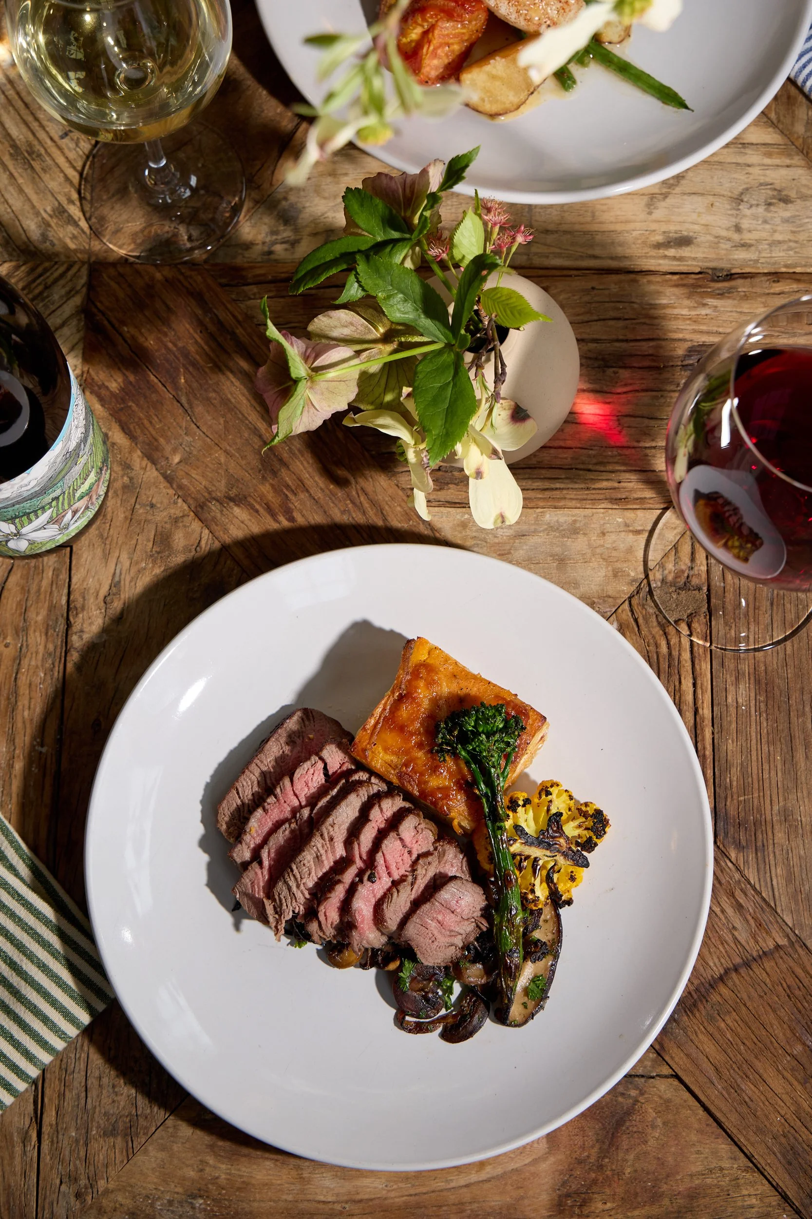 A white plate with slices of cooked steak, a slice of vegetable lasagna topped with broccoli and a grilled mushroom, on a rustic wooden table at Restaurant Foster at Lambert's Cove Inn on Martha's Vineyard. 
