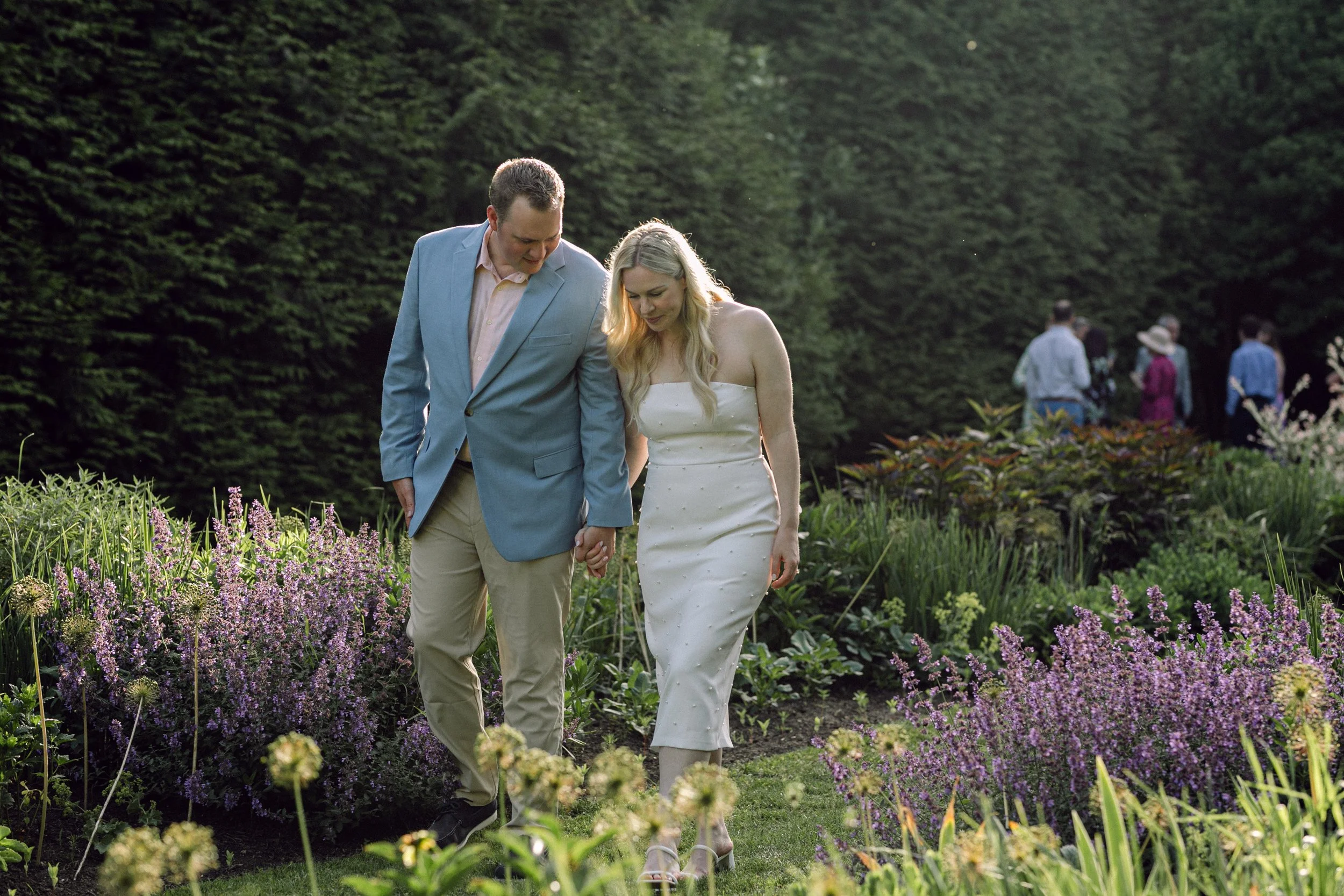 A couple holding hands walking through the English garden at Lambert's Cove Inn on Martha's Vineyard with purple flowers, with other people in the background.