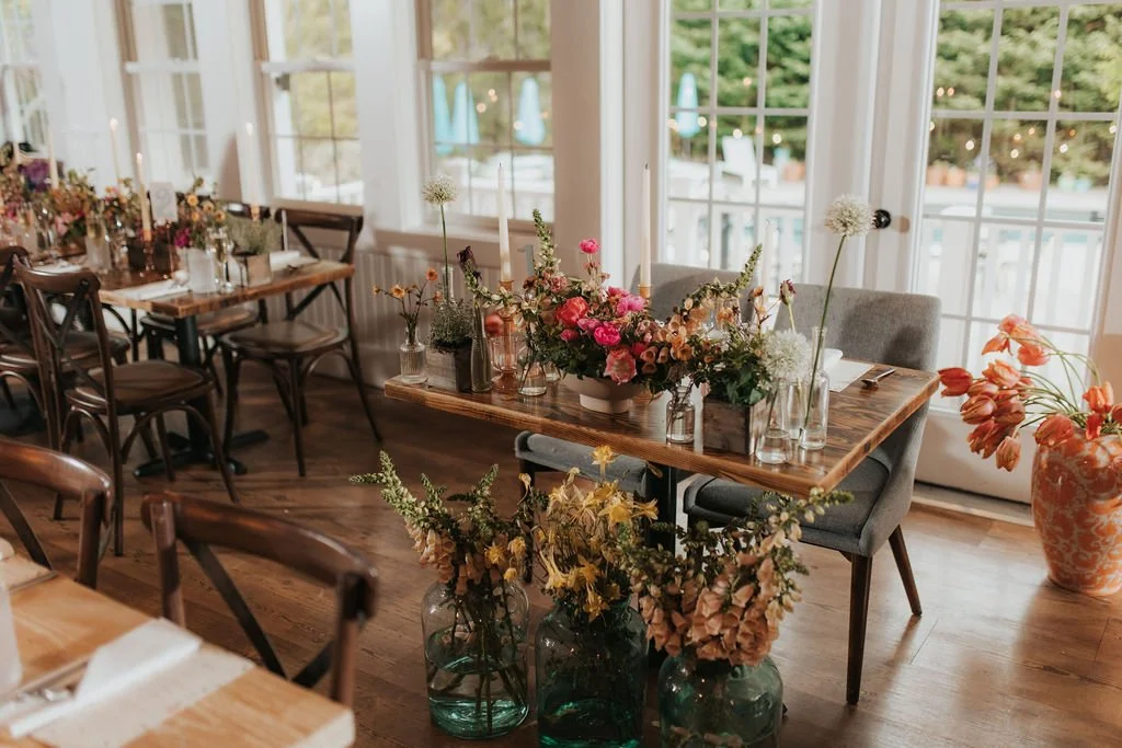 A decorated dining room with a table set for an event, featuring floral centerpieces, candles, and glassware, with large windows allowing natural light.